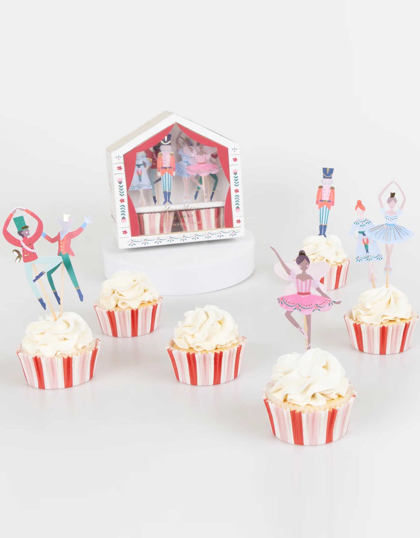 A set of cupcakes decorated with Momo Party's Nutcracker Ballet Cupcake Kit of the Nutcracker themed toppers including the Nutcracker soldier, the Mouse King, the ballerina, sugarplum fairy and its red and pink striped cupcake liners. Its stage shaped packaging box is in the background.