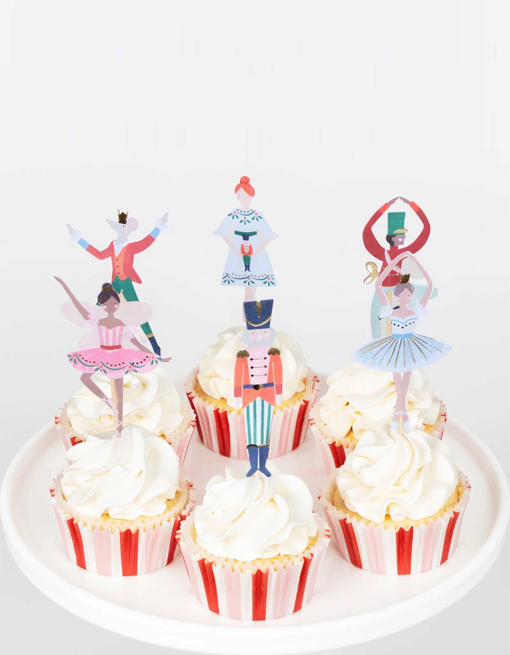 A set of cupcakes decorated with Momo Party's Nutcracker Ballet Cupcake Kit of the Nutcracker themed toppers including the Nutcracker soldier, the Mouse King, the ballerina, sugarplum fairy and its red and pink striped cupcake liners on a white cupcake stand.