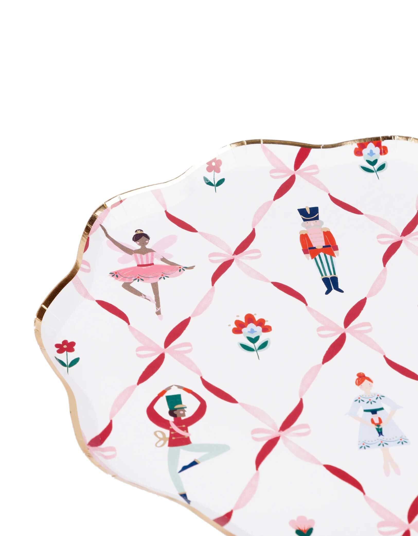 A closeup shot of Momo Party's 10.5" Nutcracker ballet dinner plate with scallop edge in gold foil with the design of pink ribbon bows, sugarplum fairies, Nutcracker soldiers, the Mouse King, ballerinas on a white background.