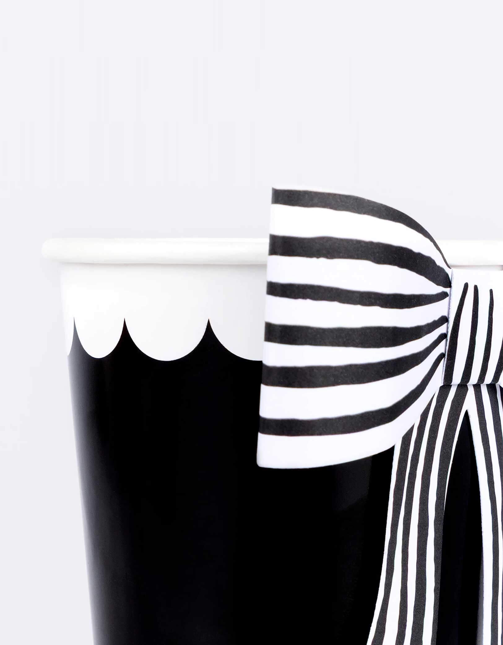 A close up shot of Momo Party's 9 oz Black & White Stripy Bow Party Cup by Meri Meri on a white background. These black cups, adorned with chic striped bows, add a spellbinding touch to your party drinks. The classic black-and-white design pairs beautifully with the rest of your Halloween tableware and decorations.