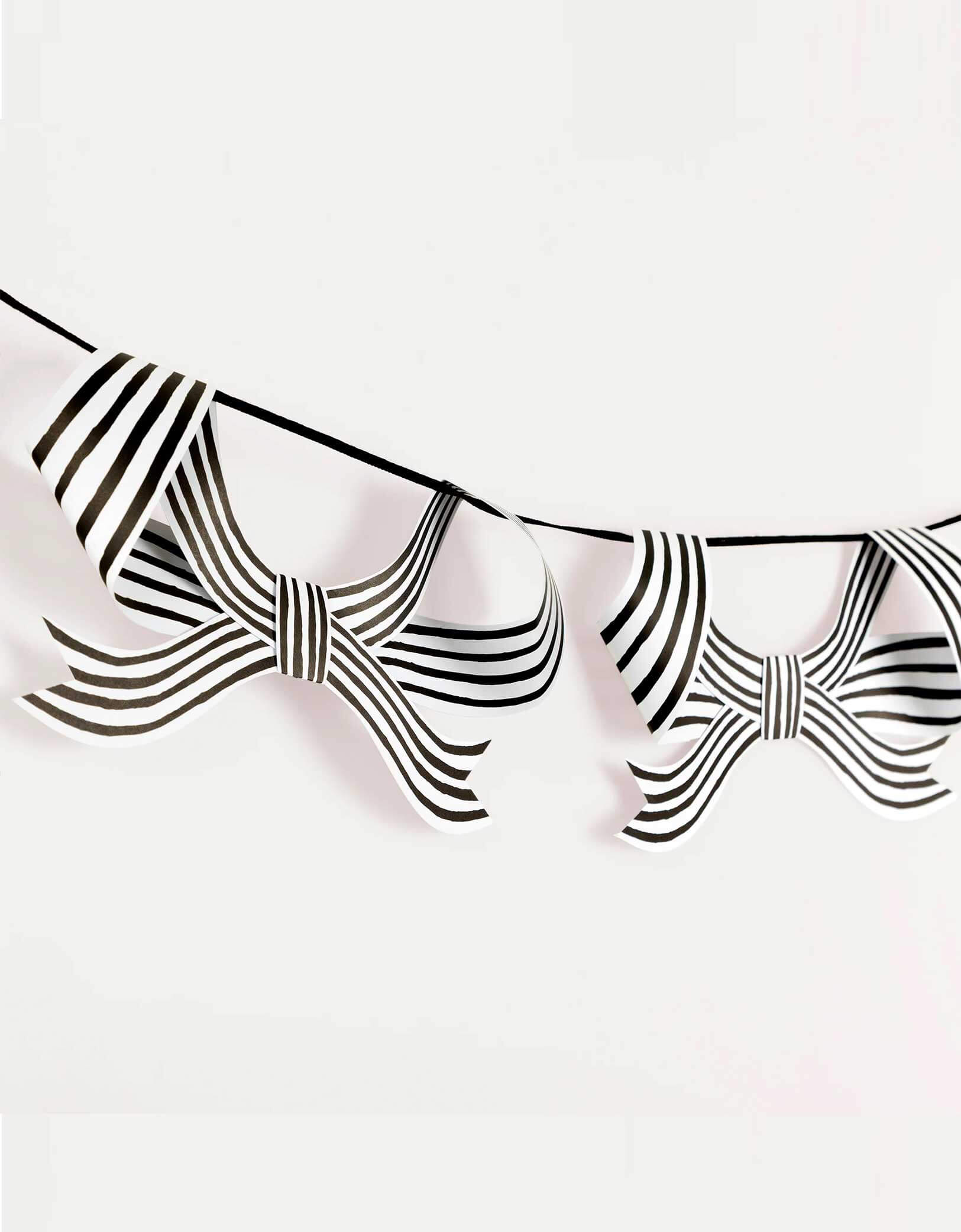 A close up shot of Momo Party's 8 ft  Black & White Stripy Bow Garland with bow-shaped ornaments by Meri Meri on a white background. The bold black and white stripes, and soft black velvet ribbon, will give a striking touch indoors or out at your Halloween celebration. Perfect for draping on the party table or mantel, hanging in the porch or windows, or swaying from trees. Plus, it's reusable to bring effortless elegance, and a hint of eerie charm, year after year.