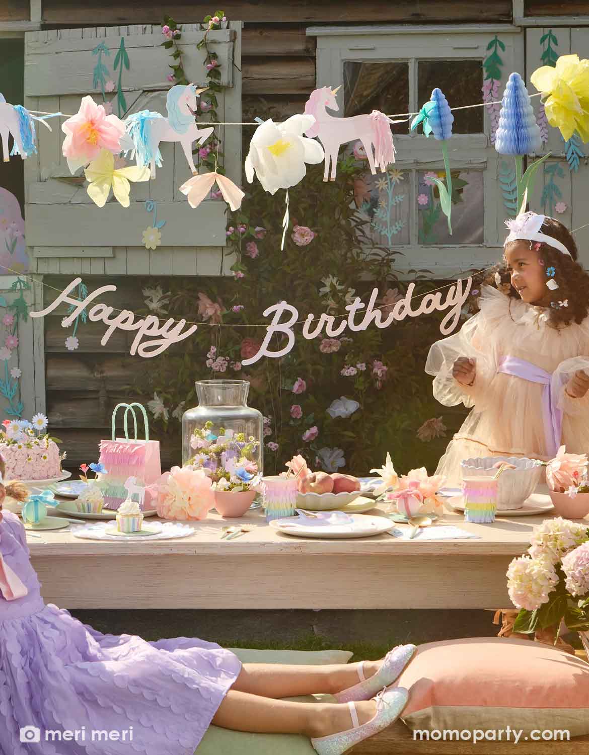 Girls with tutu dresses and unicorn headbands at a magical and whimsical unicorn themed birthday party with unicorn floral party banner with a 'Happy Birthday' sign hung above the party table which is decorated with various unicorn themed tablewares including plates, cups, napkins from Momo Party. Making this a dreamy inspo for kid's unicorn themed birthday party.