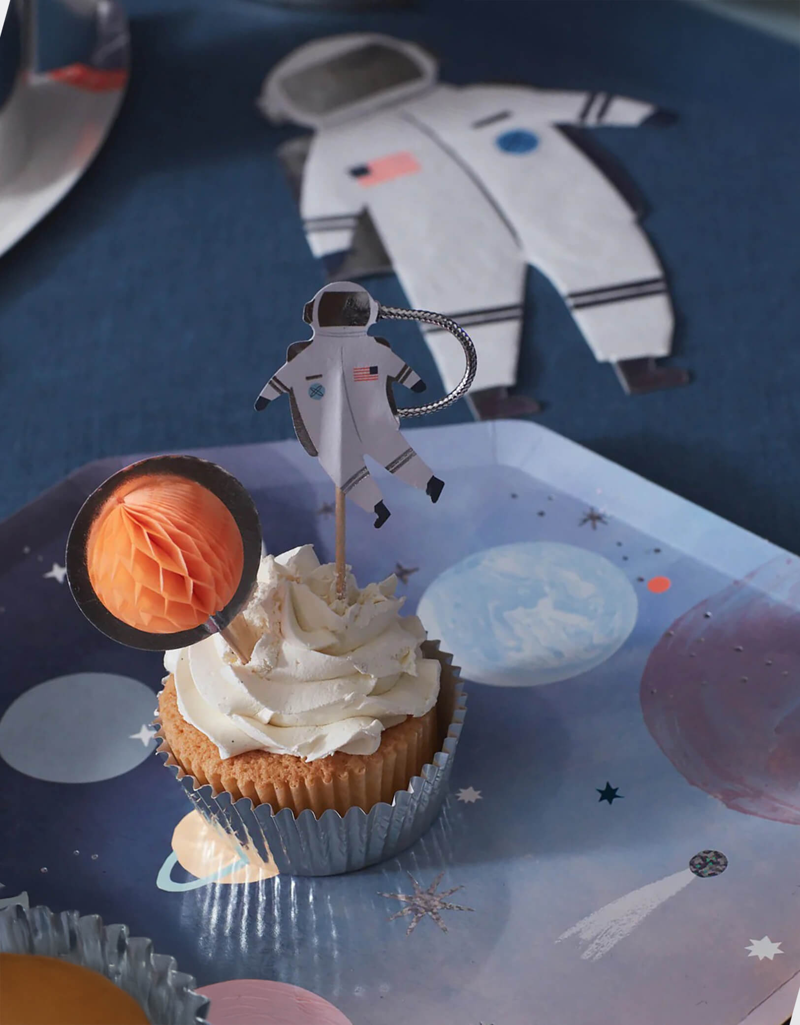 A cupcake topped with astronaut and an orange planet honeycomb topper on a space-themed dinner plate which is next to an astronaut shaped napkin on a dark galaxy themed tablecloth from Momo Party.