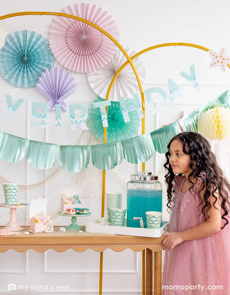 A girl in a pink tutu dress by a white wall decorated with mermaid party decorations including Momo Party's under the sea paper fans in pastel colors of aqua, pink, lilac, teal, cream and peach. Next to the paper fans hung a fishing net draping over a golden arch structure adorn with mermaid Happy Birthday teal foil banner by My Mind's Eye. The girl is standing next to a table decorated with mermaid supplies including mermaid tail party cups, under the sea treat boxes filled with treats and sugar cookies.