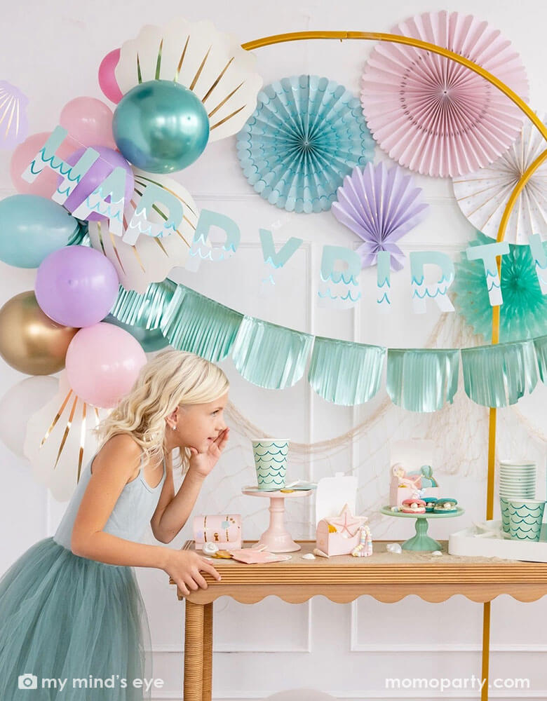 A girl in a teal tutu dress by a white wall decorated with mermaid party decorations including Momo Party's under the sea paper fans in pastel colors of aqua, pink, lilac, teal, cream and peach. Next to the paper fans hung a fishing net draping over a golden arch structure adorned with honeycomb jelly fish decorations by My Mind's Eye. She's whispering next to a table decorated with mermaid supplies including mermaid tail party cups, under the sea treat boxes filled with treats and sugar cookies.