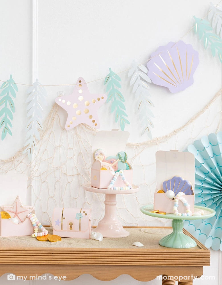 A mermaid party decoration set up on the wall features Momo Party's under the sea paper fan set in pastel colors aand the jumbo mermaid garland featuring seagrass, sea stars and sea shells in dreamy pastel colors by My Mind's Eye, underneath hung a fishing net draping over the party table which has multiple treasure box shaped treat boxes filled with treats and mermaid themed sugar cookies, making this an perfect inspo for an enchanting kid's mermaid themed birthday party set up.