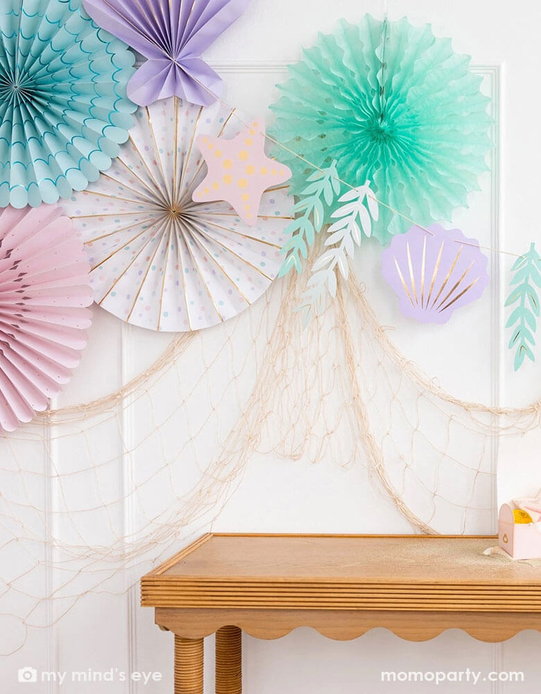 A mermaid party decoration set up on the wall features Momo Party's under the sea paper fan set which includes a set of 6 paper fans 6 fans of size 14", 8.5", 8", 7", and 5.5" in pastel colors of aqua, pink, lilac, teal, cream and peach. Next to the paper fans hung the jumbo mermaid garland by My Mind's Eye in the matching colors, underneath hung a fishing net draping over the party table, making this an perfect inspo for an enchanting kid's mermaid themed birthday party set up.