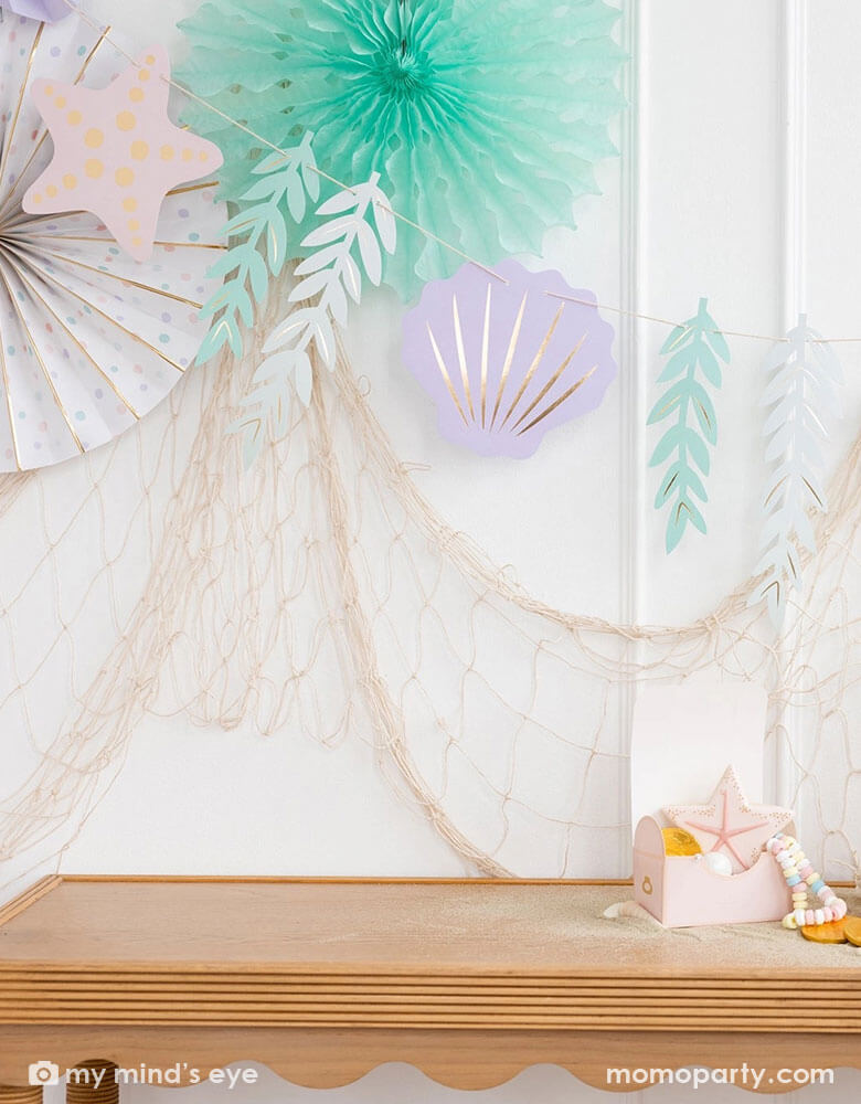 A mermaid party decoration set up on the wall features Momo Party's under the sea paper fan set which includes a set of 6 paper fans 6 fans of size 14", 8.5", 8", 7", and 5.5" in pastel colors of aqua, pink, lilac, teal, cream and peach. Next to the paper fans hung the jumbo mermaid garland by My Mind's Eye in the matching colors, underneath hung a fishing net draping over the party table, making this an perfect inspo for an enchanting kid's mermaid themed birthday party set up.