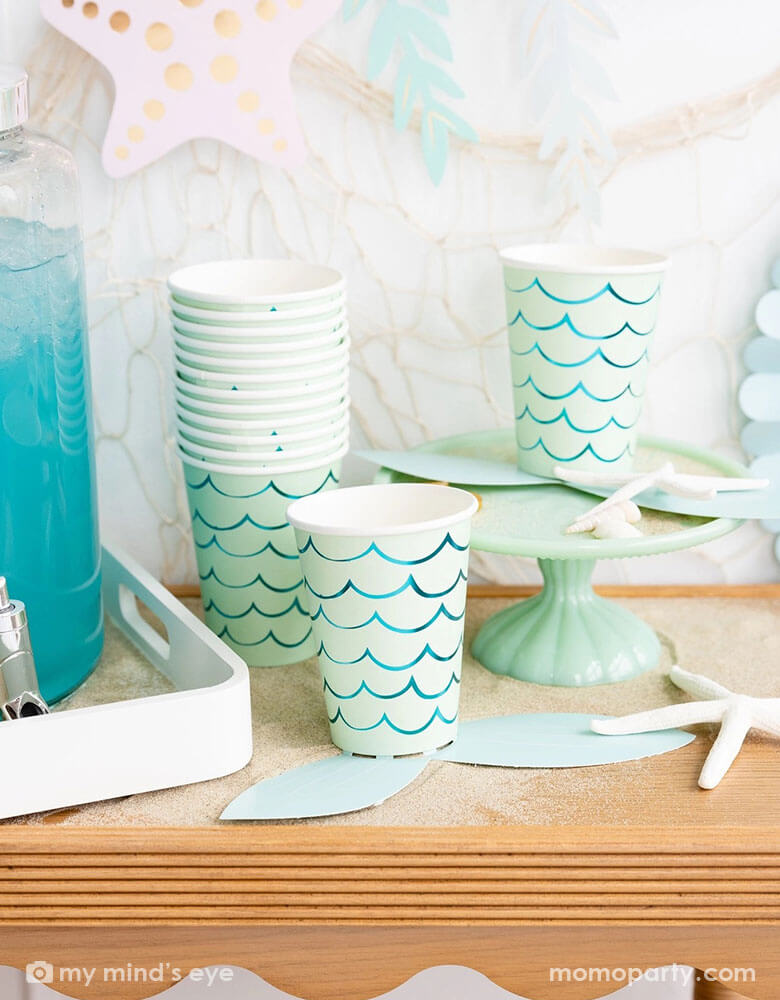 A mermaid party table features Momo Party's mermaid tail paper cups by My Mind's Eye. Next to the paper cups is a water dispenser filled with aqua colored drink. The table is decorated with shells, sea star and white sand decorations. In the back there's a fishing net hung on the wall which is adorned with under the sea paper party pennants in soft pastel colors, making this a perfect inspo for a kid's mermaid, under the sea party decorations. 