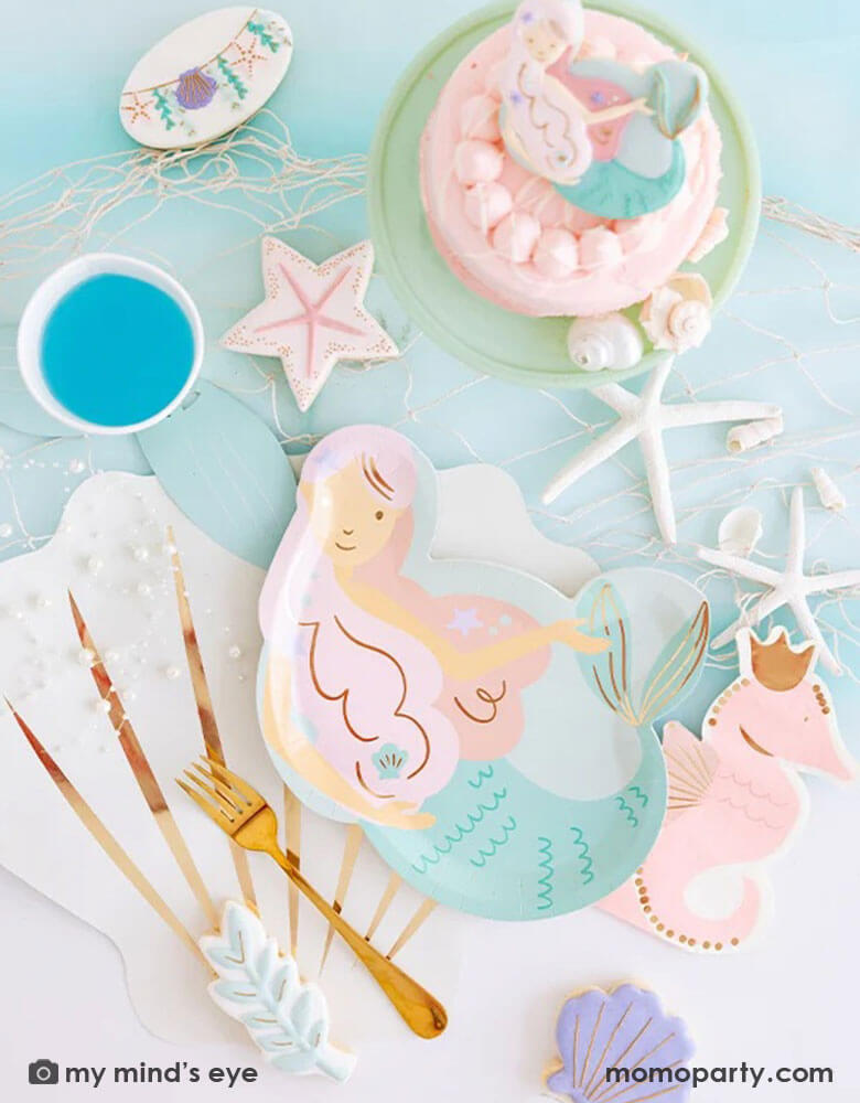 An enchanting mermaid party tablescape features Momo Party's mermaid shaped plate, pink seahorse shaped napkin, and mermaid tail party cup on a cream seashell shaped placemat by My Mind's Eye. Around these under the sea inspired tableware, there are mermaid themed sugar cookies and a small pink cake, along with some sea star decorations and fishing net, this makes a perfect inspo for kid's magical mermaid party ideas.