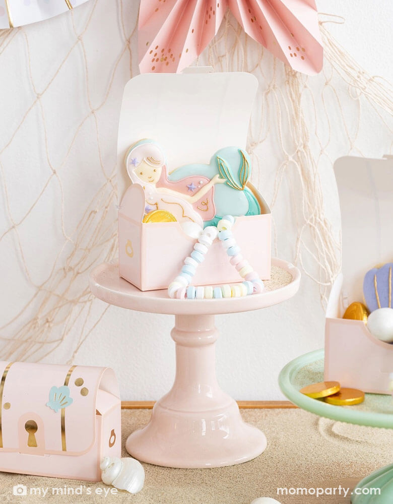 A closeup shot of an adorable mermaid party table decorated with Momo Party's pink under the sea treasure chest treat boxes on darling pastel cake stands. In the treat boxes are pastel candy necklaces, some gold chocolate coins, mermaid and seashell sugar cookies in coordinating pastel colors. In the back of the wall there's a fishing net decoration draped from the wall, making this a great decorating inspo for kid's mermaid/under the sea birthday parties.