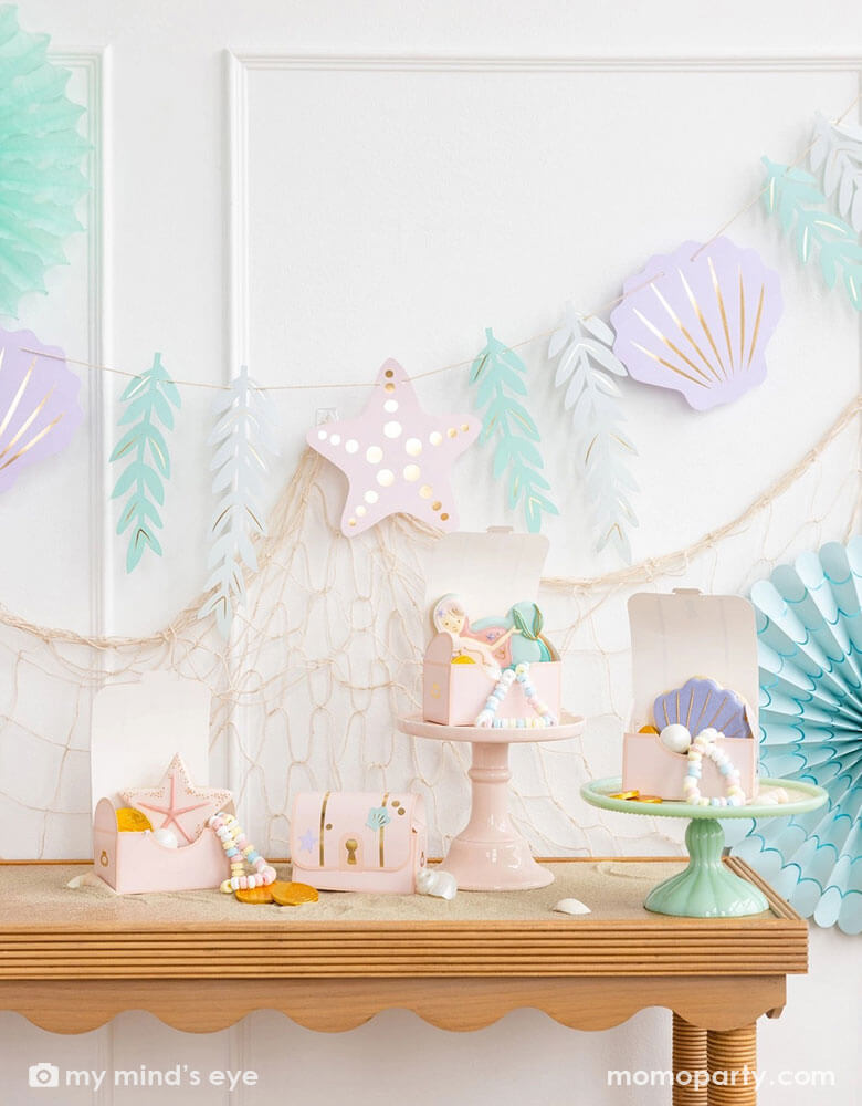 A mermaid party decoration set up on the wall features Momo Party's under the sea paper fan set in pastel colors aand the jumbo mermaid garland featuring seagrass, sea stars and sea shells in dreamy pastel colors by My Mind's Eye, underneath hung a fishing net draping over the party table which has multiple treasure box shaped treat boxes filled with treats and mermaid themed sugar cookies, making this an perfect inspo for an enchanting kid's mermaid themed birthday party set up.