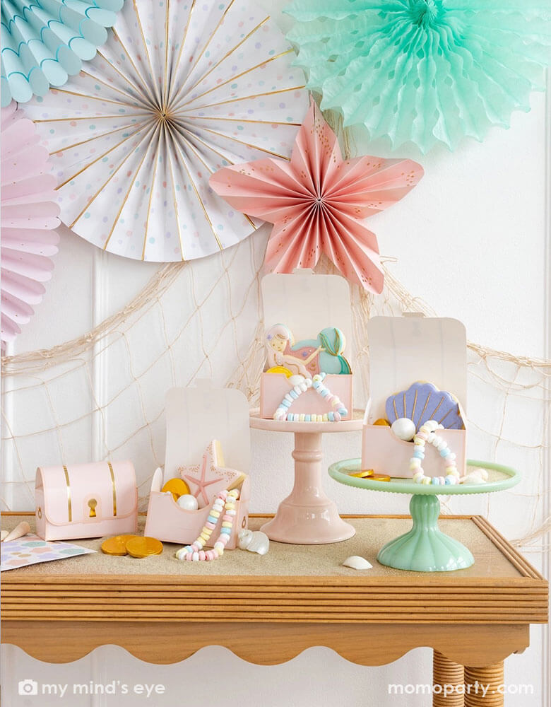A mermaid party table with Momo Party's pink under the sea treasure chest treat boxes by My Mind's Eye. In these treat boxes are some pastel colored treats, mermaid themed sugar cookies with designs of a mermaid, a seashell and a sea star in soft pastel colors. Some of them are placed on pastel colored cake stands. The wall behind is decorated with Momo Party's under the sea paper fan set in pastel colors of aqua, pink, lilac, teal, cream and peach, with a fishing next hung underneath.