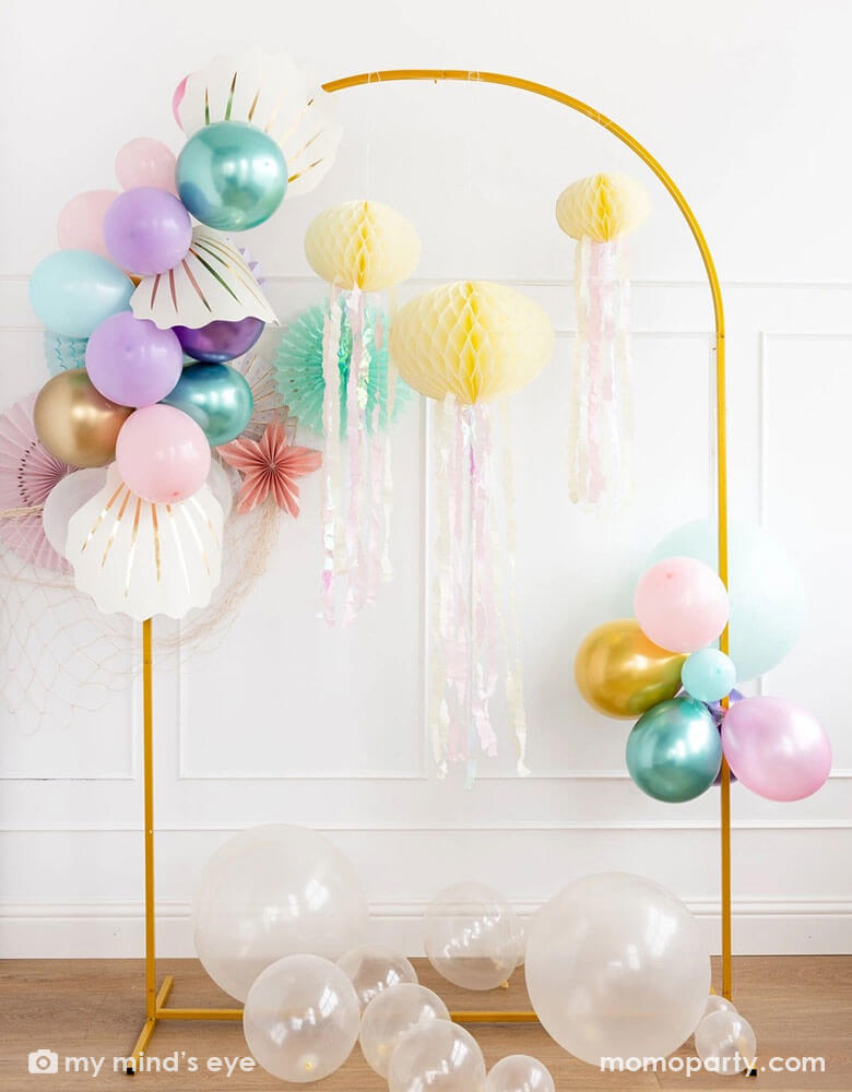 A white wall decorated with mermaid party decorations including Momo Party's under the sea paper fans in pastel colors. Next to the paper fans hung a fishing net draping over a golden arch structure adorned with honeycomb jelly fish decorations by My Mind's Eye. On the left side there is a mermaid inspired balloon garland adorned with shell shaped paper decorations in cream. On the floor there are some clear latex balloons as bubbles, making this an inspo for an enchanting kid's mermaid party set up.