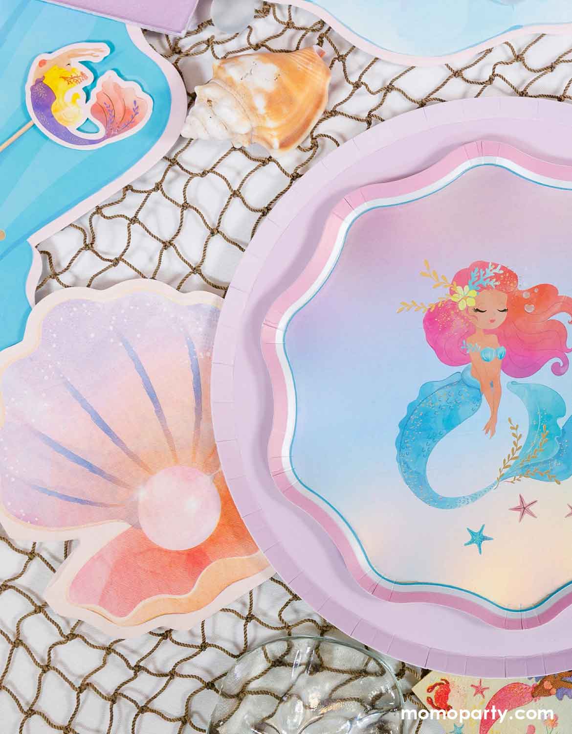 A kid's mermaid-themed party table featuring various under the sea themed party tablewares from Momo Party including paper plates with a colorful mermaid design, pastel shell shaped napkin, blue shell shaped party placemats and mermaid cupcake toppers, along with the fishing net underneath, it makes a whimsical tablescape for kid's mermaid themed birthday parties.