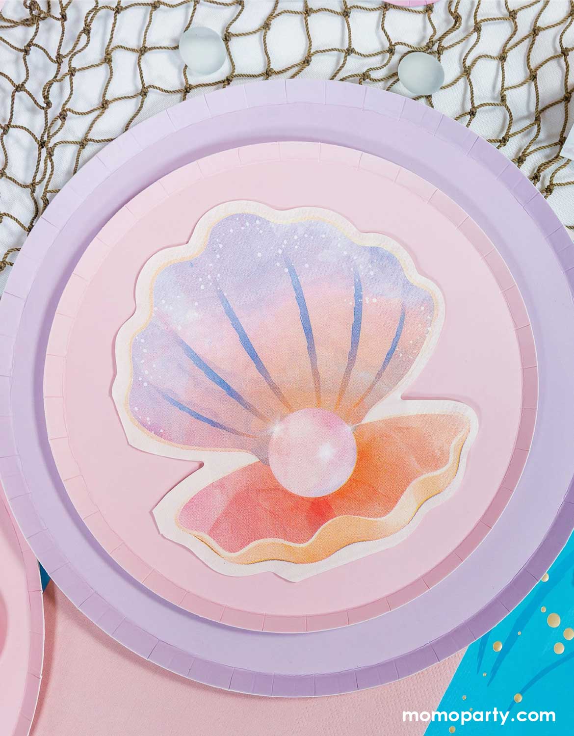 A mermaid themed tablescape featuring Momo Party's pink and lilac paper plates with a shell shaped napkin on a fishing net surface.