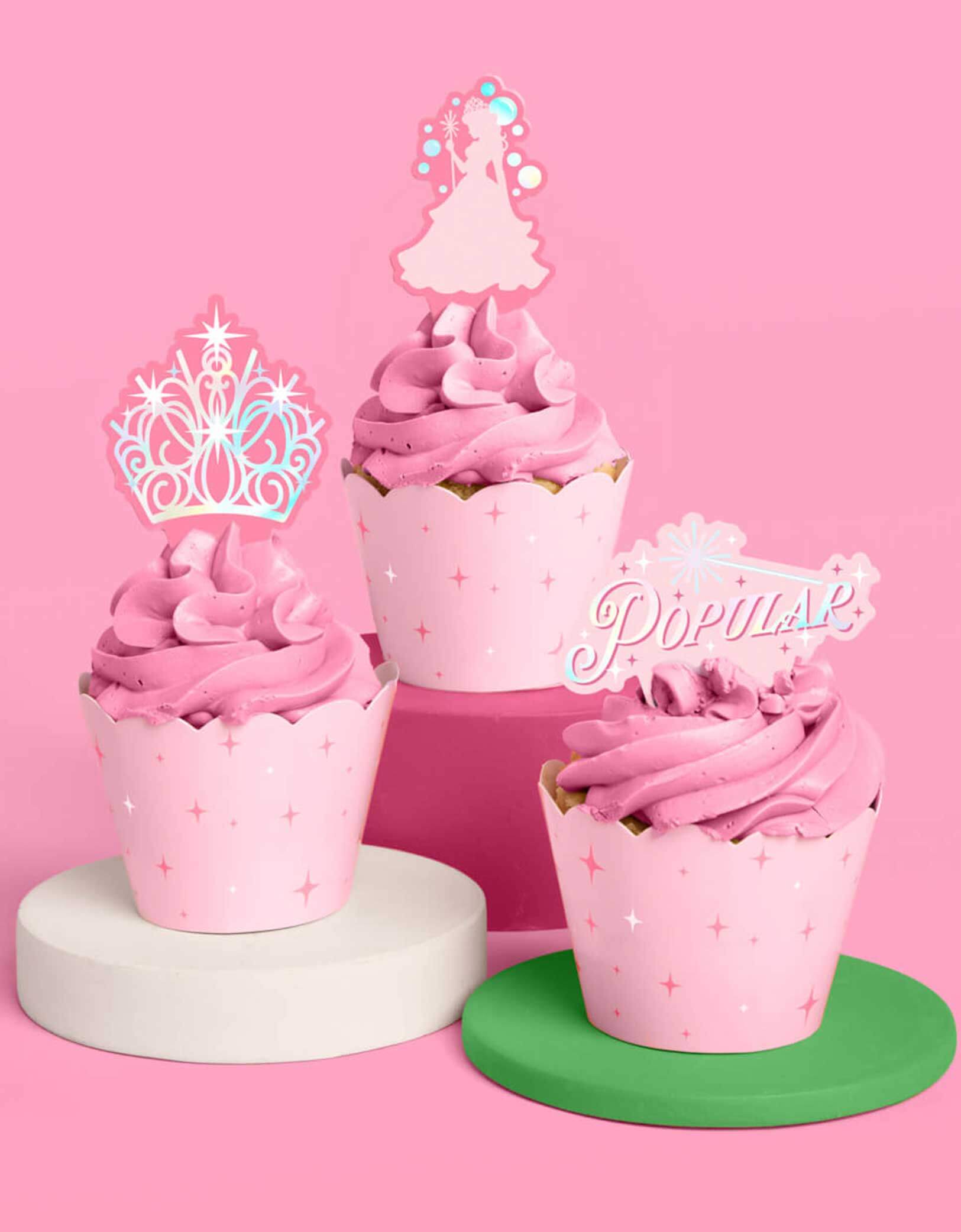 Three pink good Wicked themed cupcakes wrapped with spellbinding green liners with witch inspired star point design on the side with good witch-themed toppers including the designs of Glinda, a tiara and Popular text with a wand from Momo Party on a pink background.