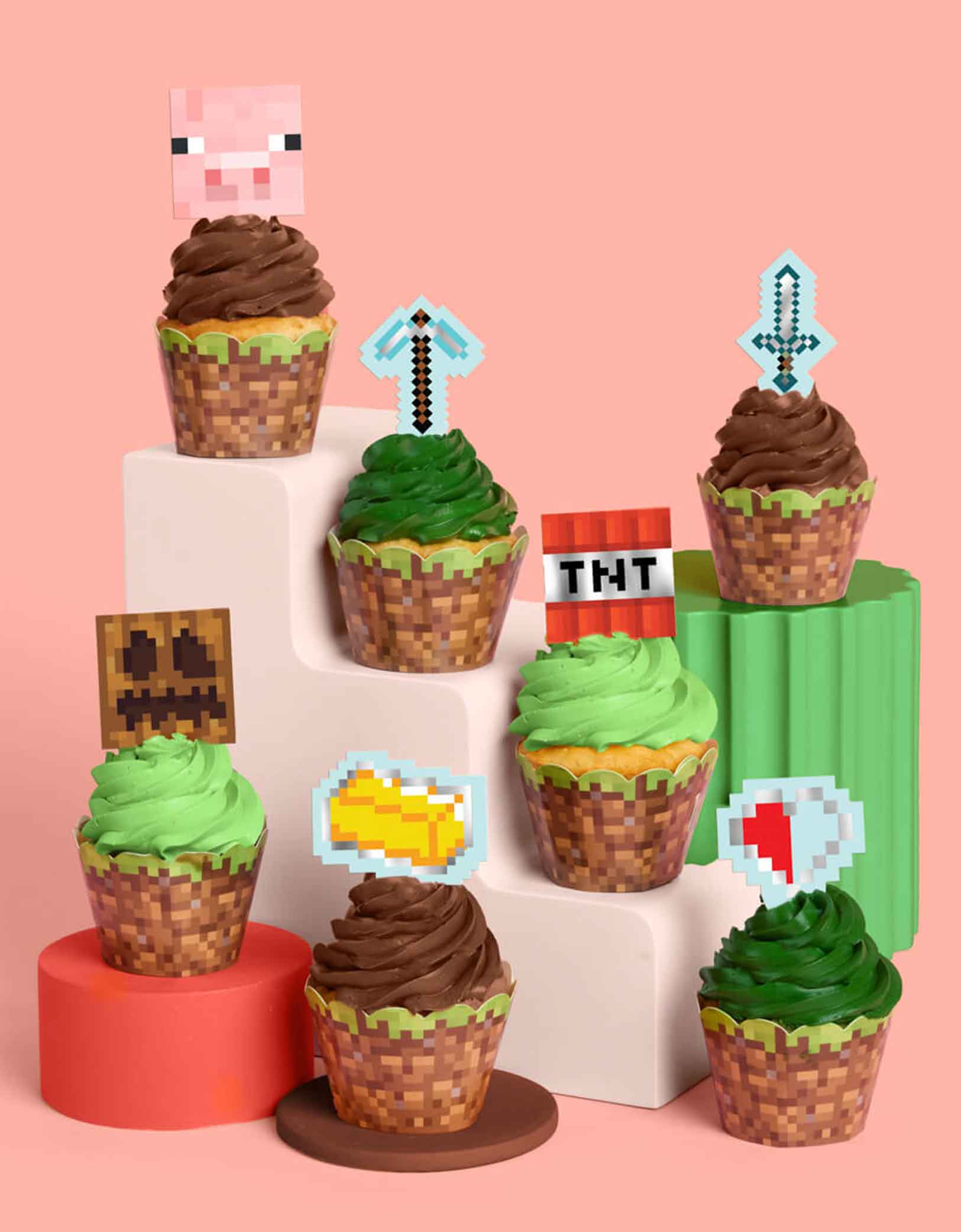 A set of Minecraft-themed cupcakes in green and brown with decorative Minecraft themed toppers and liners featuring pig face, pixel swords, TNT, gold bricks etc from Momo Party on a pink background.