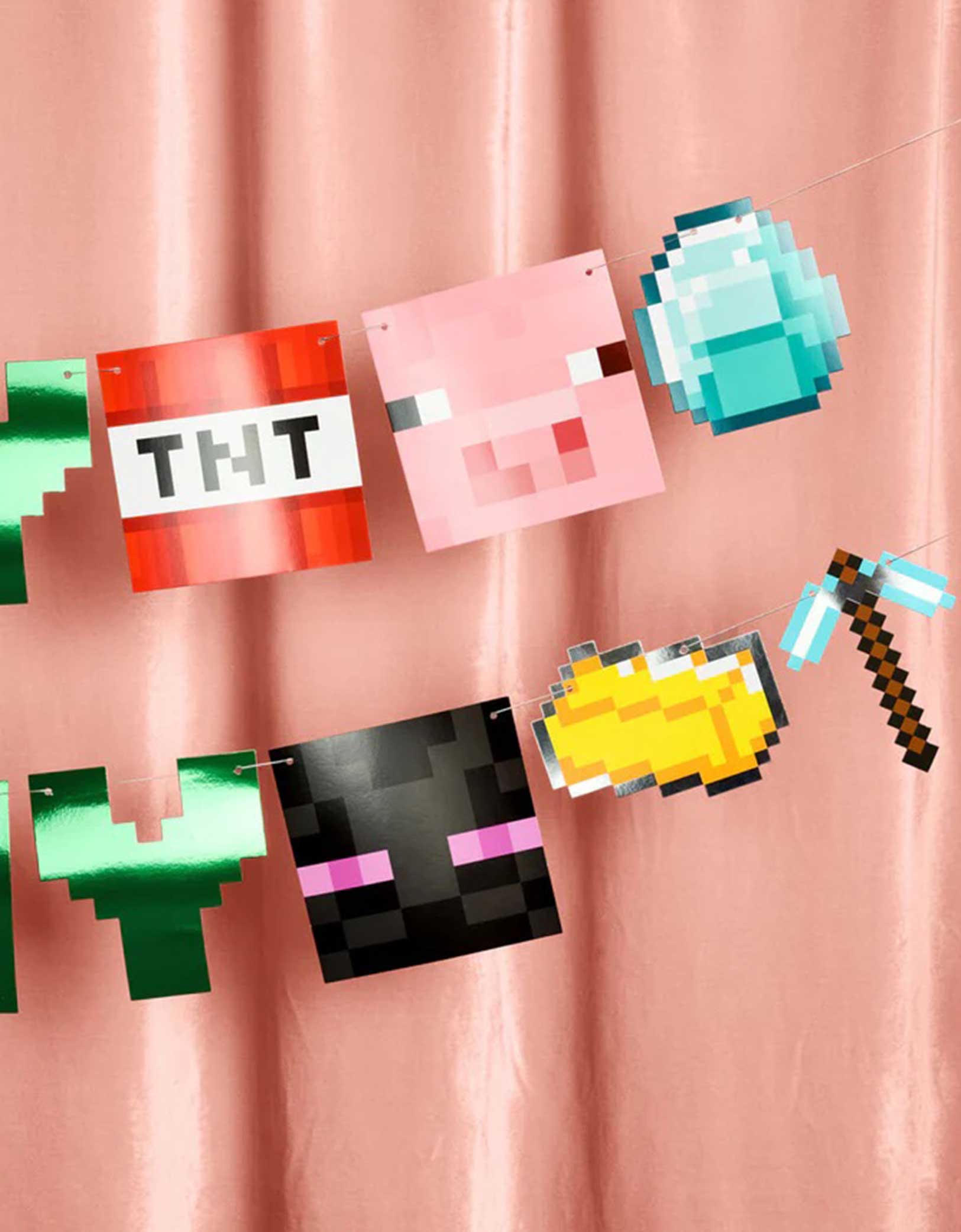 A closeup shot of Momo Party's Minecraft Pixel Birthday Banner with pixelated pennants including TNT, a pink pig block, a diamond block, and other Minecraft-style blocks on a pink curtain background.