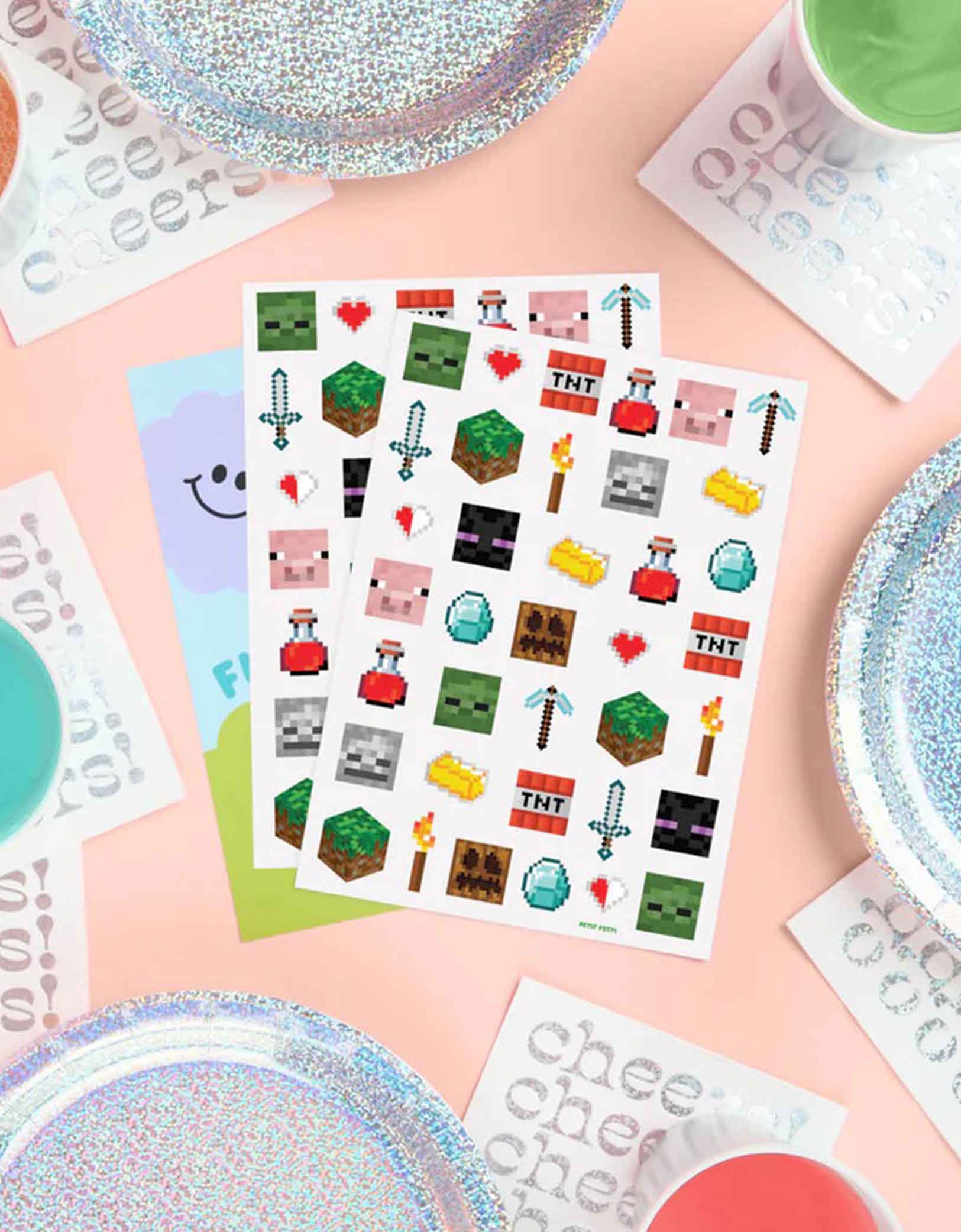 Two sheets of Minecraft-themed temporary tattoos featuring pixelated block designs including TNT, creeper, pig, pixel axes, pixel swords etc. from Momo Party on a pink surface with colorful plates.