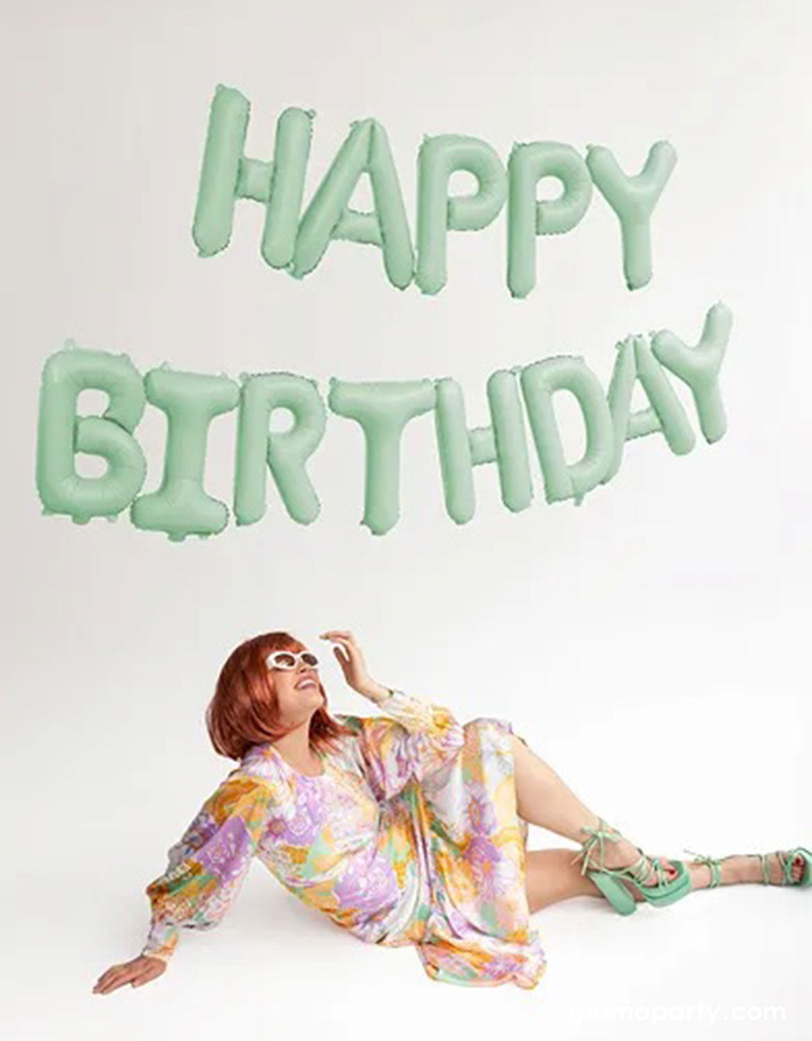 A woman in a floral dress and sunglasses sitting on the floor with Momo Party's 14" mint 'Happy Birthday' balloon set banner above her on a white background.