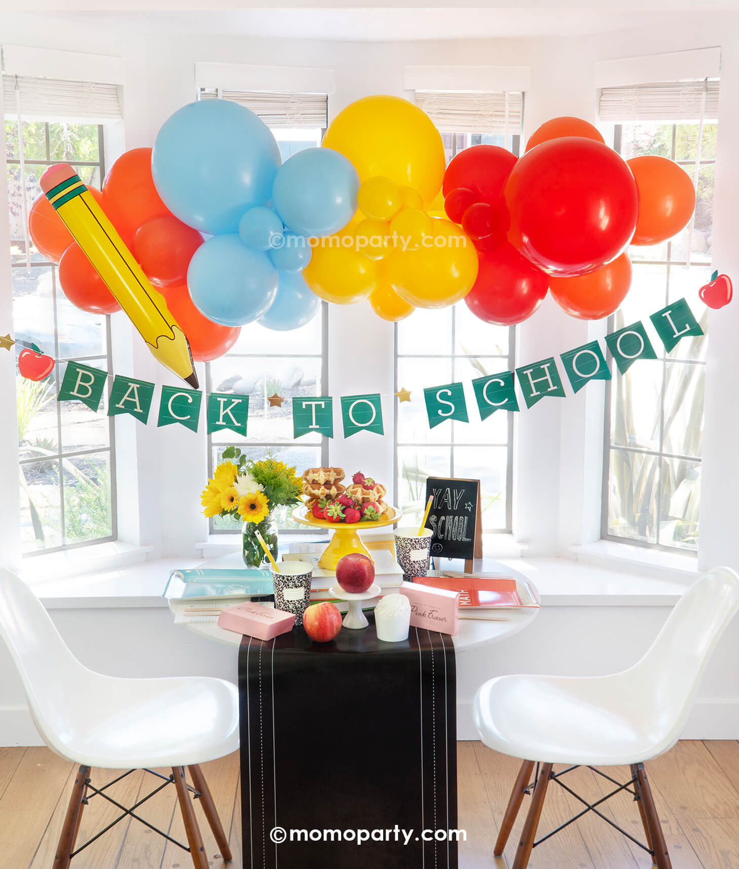 Momo Party's School Day Party Box set up featuring multiple school themed party goods including the back to school apple party banner, a festive balloon garland in the school inspired colors adorned with a pencil inflatable. On the table are text book shaped plates in different colors, eraser shaped treat boxes, notebook party cups and pencil straws on the chalkboard table runner. With stacked waffles, treats and fruits, this is a perfect idea for your child's first day of school breakfast celebration!