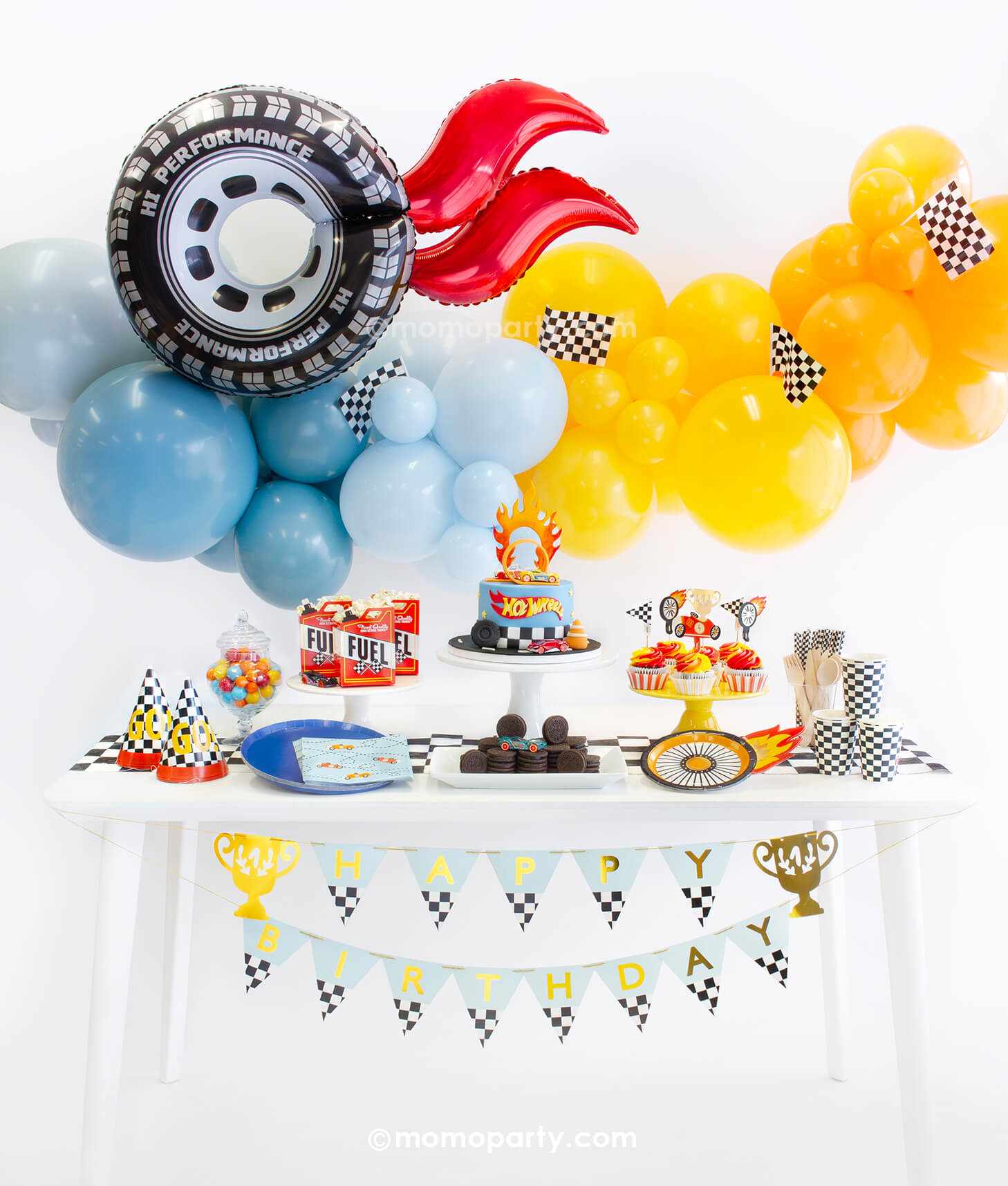 Momo Party's Hot Wheel Party Box featuring a orange and blue organic balloon garland topped with race car checkered flags and wheel shaped foil balloon with red flame. On the table there are wheel shaped plates, midnight blue dinner plates, race car blue napkins, checkered party cups, birthday hats and cupcake topped with race car themed toppers. In the middle is a fondant Hot Wheels birthday cake, along with the trophy HBD banner, a perfect party inspiration for boy's Hot Wheels themed birthday party.