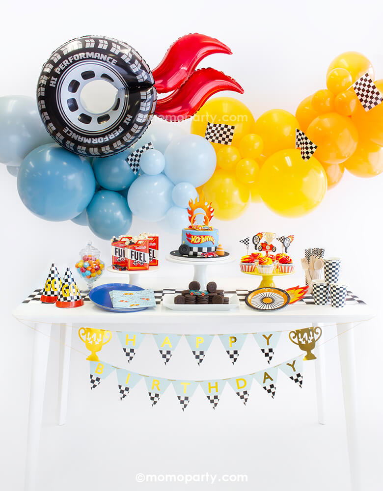 Momo Party's Hot Wheel Party Look, featuring a orange and blue organic balloon garland topped with race car checkered flags and wheel shaped foil balloon with red flame. On the table there are wheel shaped plates, midnight blue dinner plates, race car blue napkins, checkered party cups, birthday hats and cupcake topped with race car themed toppers. In the middle is a fondant Hot Wheels birthday cake, along with the trophy HBD banner, a perfect party inspiration for boy's Hot Wheels themed birthday party.