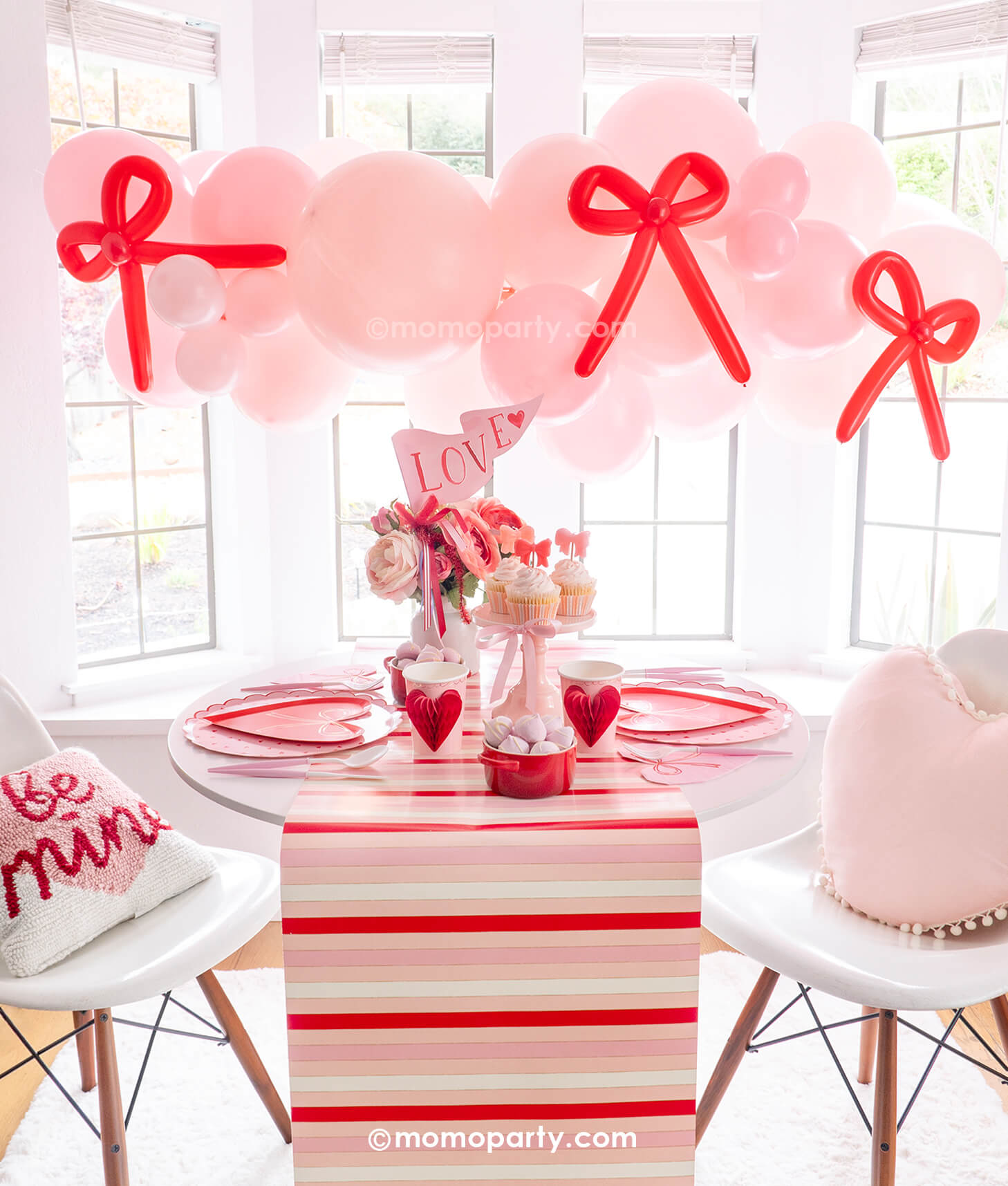 A beautiful party set up next to a big, bright window featuring Momo Party's Bow-tiful Valentine's Day Party Box for 8 guests. Featuring a pink balloon garland with red bow balloons on it, heart shaped tableware including plates, napkins and party cups, and a beautiful flower bouquet with a pink "LOVE" party pennant, this adorable party box in classic Valentine's colors of red and pink is perfect for your Valentine's Day party or Galentine's Day celebration.