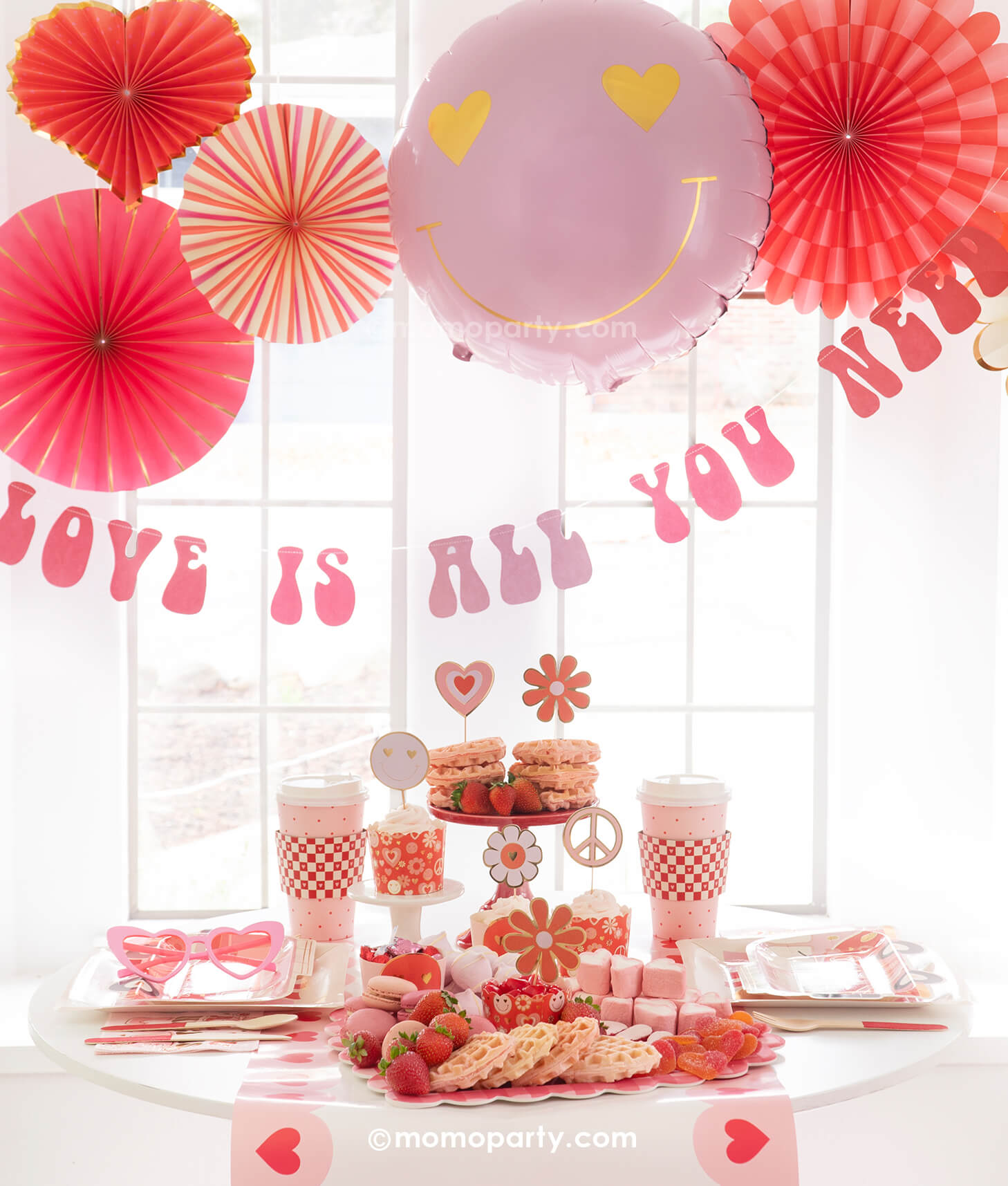 A Valentine's Day celebration featuring Momo Party's Groovy Valentine's Day Party box set up with pink and red paper fans in heart shape and a "love is all you need" banner in retro type hung below the paper fans. On the table you can see a sweet board featuring treats and snacks in red and pink colors including heart shaped waffles, strawberries, macarons, marshmallows etc. Next to the desert board featured retro inspired Valentine's plates, party cups and napkins,  a perfect inspo for Vday celebration!