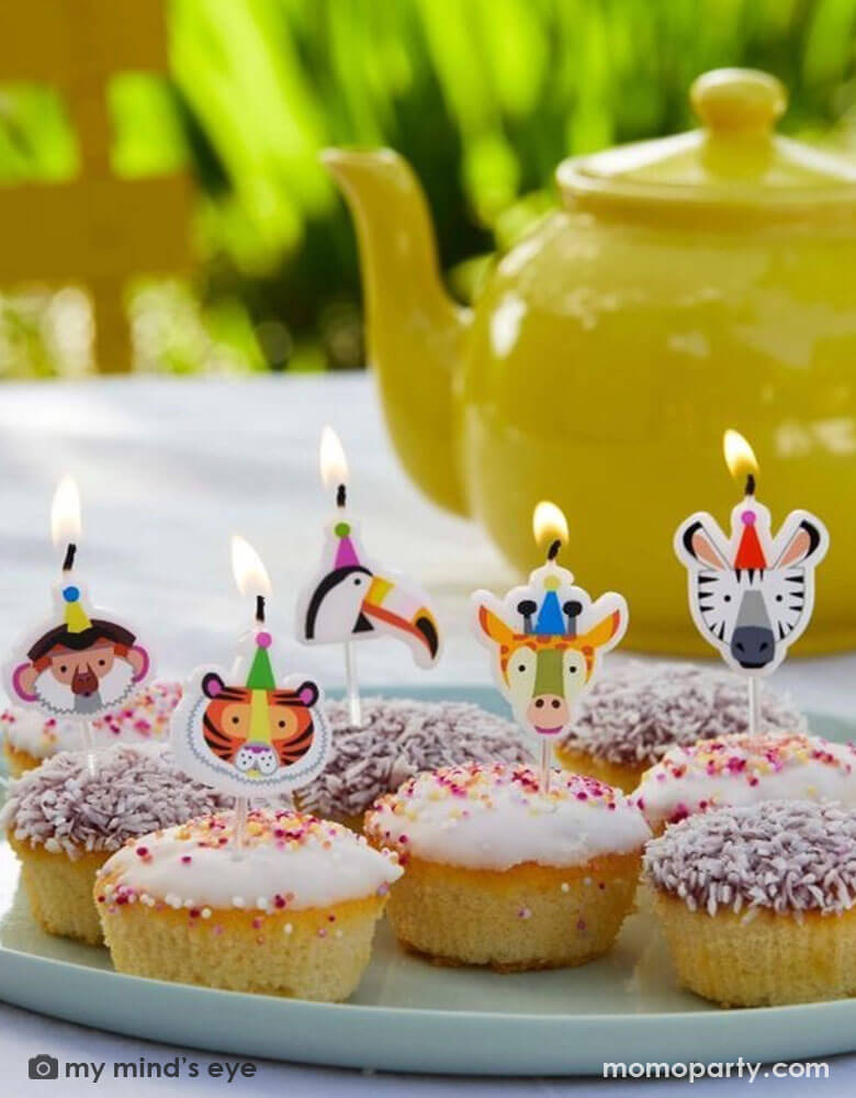 A few birthday cupcakes topped with Momo Party's animal birthday candles of a tiger, a zebra, a monkey, a toucan and a giraffe by Talking Tables. In the back is a yellow tea pot on the table ready for an afternoon time party.