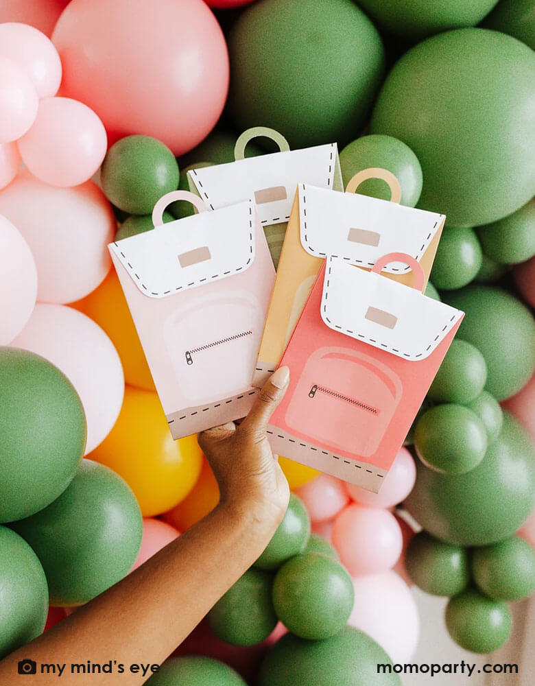 A mom holding a stack of Momo Party's backpack inspired party treat boxes in four colors by My Mind's Eye in front of a colorful balloon wall.