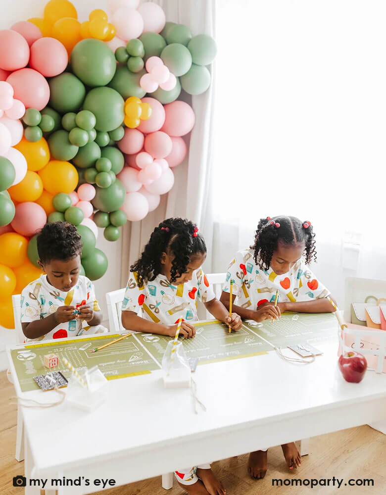 A kid's back to school first day of school celebration set up featuring a colorful balloon wall in the back with three school aged kids playing word games on Momo Party's back to school placemats by My Mind'd Eye. Each of the kid is wearing cute back to school pajamas, makes it a great idea to celebrate the first day of school with your kids.
