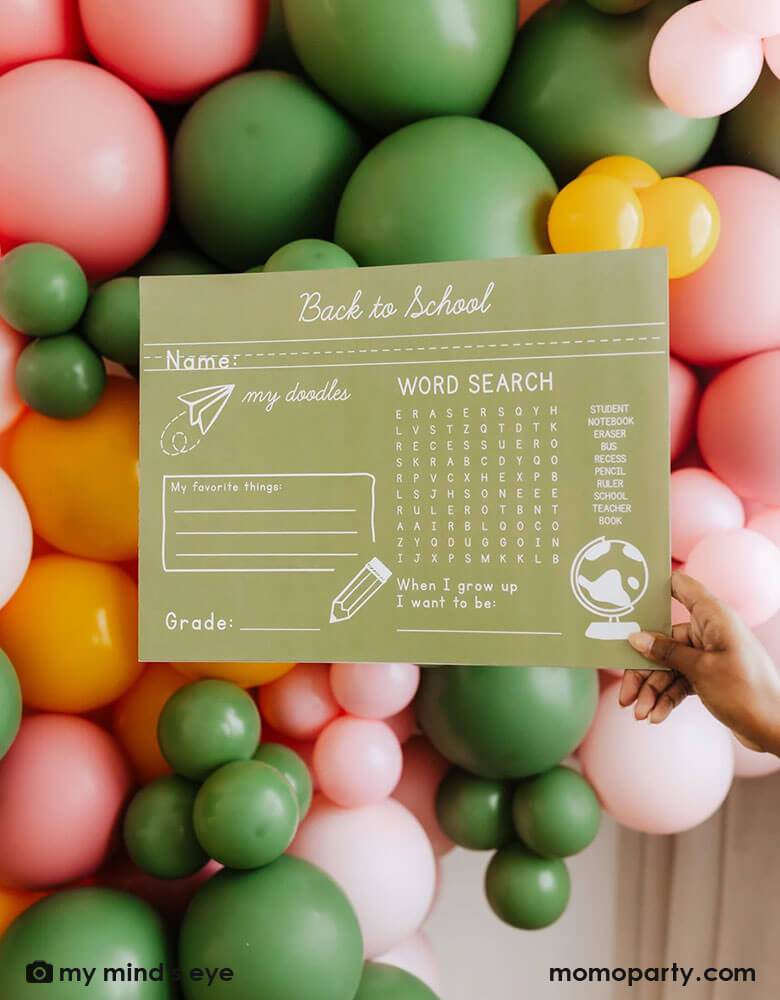 A mom holding Momo Party's 15.5 x 11.5 back to school placemat in front of a colorful balloon wall to celebrate kid's first day of school.