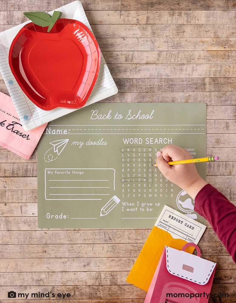 A kid holding a pencil playing work search game on Momo Party's back to school paper tablemat by My Mind's Eye. Around the tablemat, there are apple shaped plates layered with notebook square plates and pink eraser shaped napkins, at the right bottom corner of the photo, there are a report card shaped napkin with backpack shaped treat box. All in all makes a great inspiration for kid's back to school celebration.