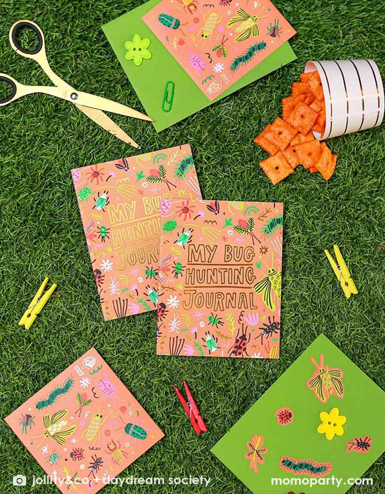 Two kid's bug hunting journals and Momo Party's backyard bug sticker sheets with some crafting tools laying on a lawn, with a foodcup filled with cheez-it crackers, makes it a cool party activity for a kid's bug themed birthday celebration or playdate.