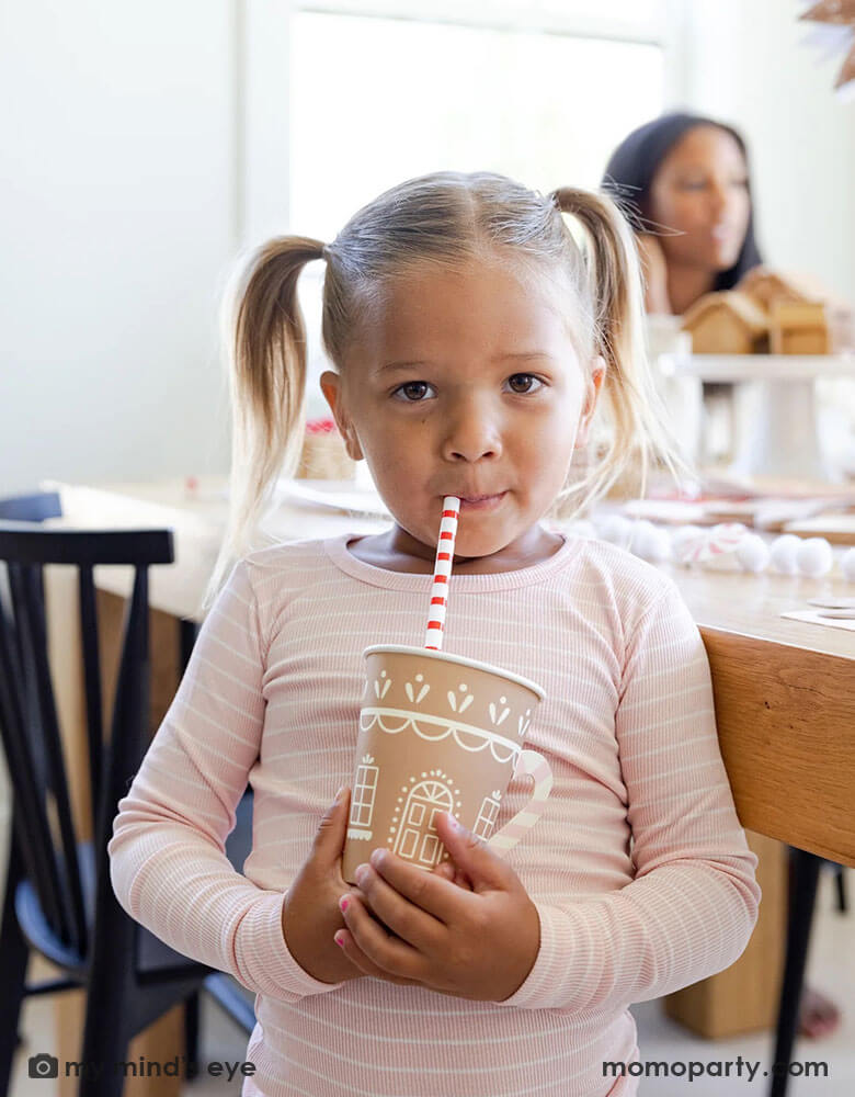 In pink-striped PJs, a girl savors a sip through Gingerbread Reusable Straws, cradling Gingerbread House Paper Party Cups with Handles. In front of a wooden dinner table, she's surrounded by captivating gingerbread-themed party essentials, all available at momoparty.com