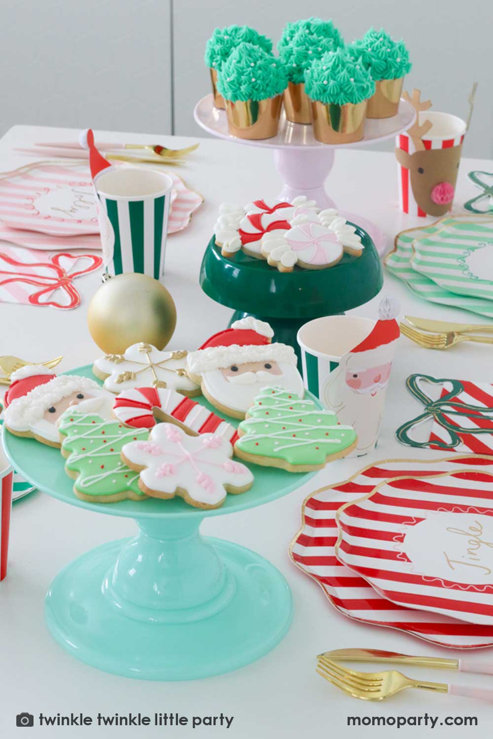 A festive Christmas party table featuring Momo Party's Festive striped dinner plates, side plates, present with a bow shaped napkins and Christmas honeycomb party cups in Santa design by Meri Meri. In the center of the table are a mint and green cake stand with festive Holiday cookies in the design of Peppermint, Santa, Christmas tree, candy cane and snowflake. Makes this a modern inspo for a festive Holiday table setting.