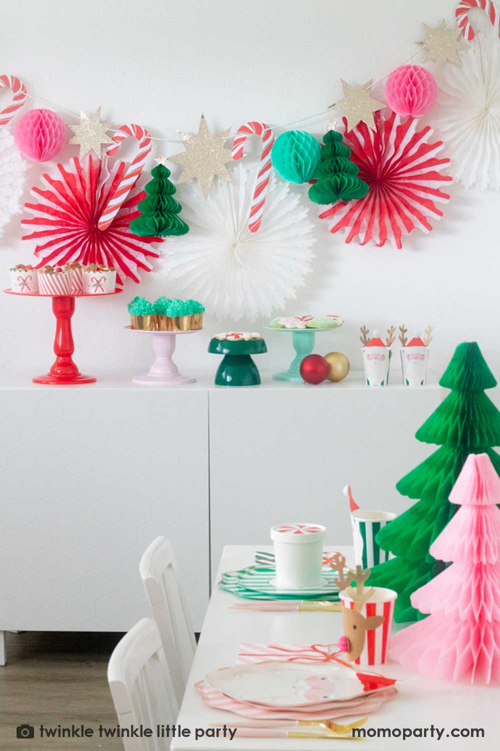 A Christmas party table featuring Momo Party's festive striped party tableware in pink red and green with Christmas tree honeycombs as the centerpiece. In the back is a Christmas dessert table decorated with Momo Party's Christmas honeycomb decorations featuring candy canes, honeycomb Christmas trees and mint honeycomb balls. On the table there are multiple cake stands in red, pink, green and mint with Holiday treats like cookies, cupcakes and baked food, making this a perfect Holiday party inspo.