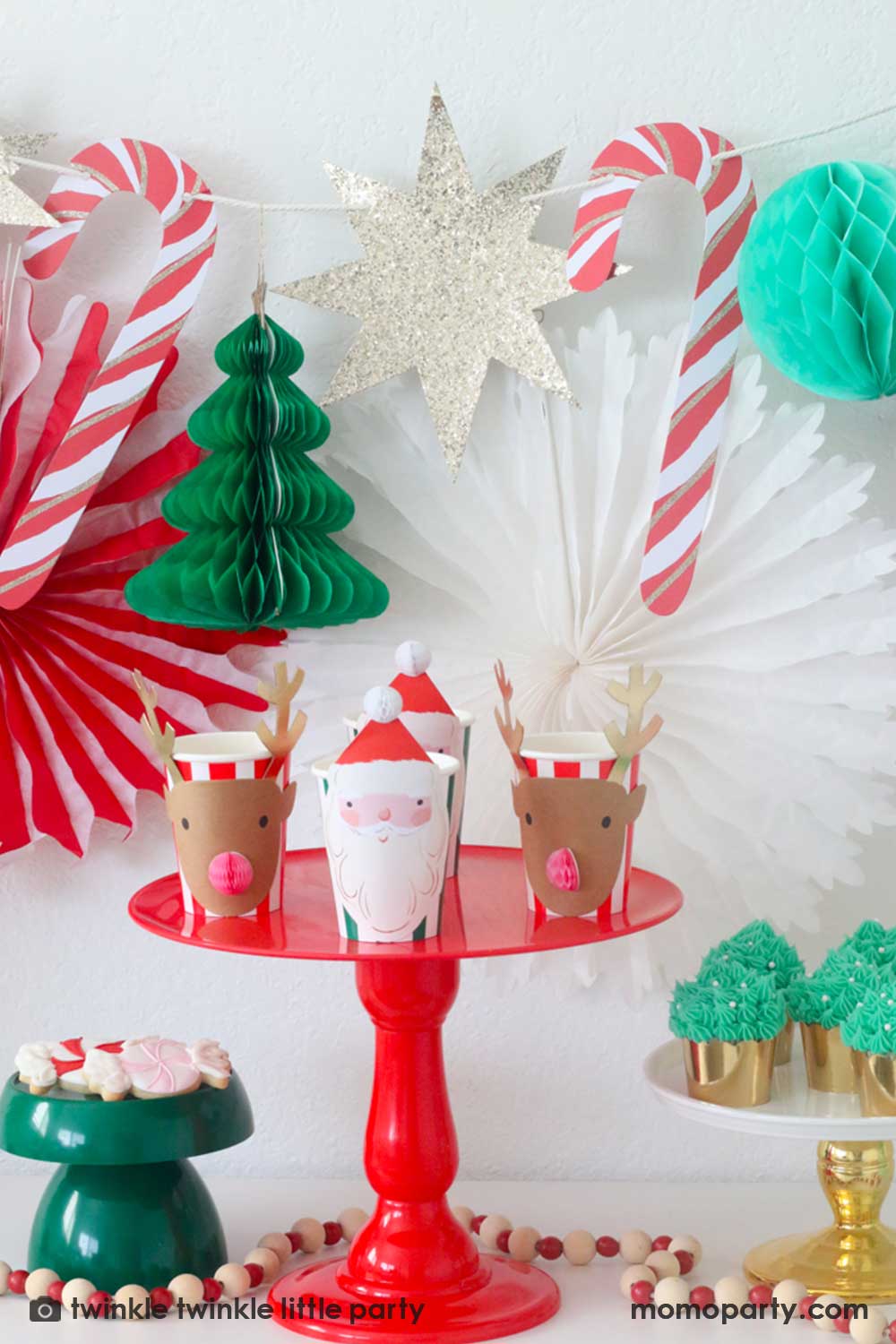 A Christmas Dessert Table decorated with Momo Party's Christmas honeycomb decorations featuring candy canes, honeycomb Christmas trees and mint honeycomb balls. On the table there's a red cake stand with Meri Meri's Christmas honeycomb party cups in the design of Santa and reindeer, and a gold cake stand with Christmas tree buttercream cupcakes and a small dark green cake stand with peppermint sugar cookies, making this a perfect  inspo for a festive Holiday party setup.