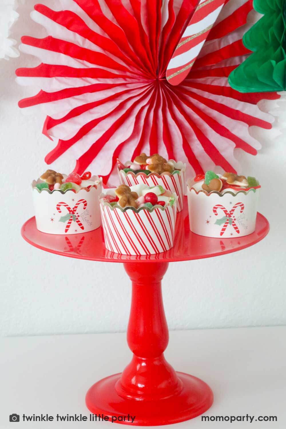 A Christmas Dessert Table decorated with Momo Party's Christmas honeycomb decorations featuring candy canes, honeycomb Christmas trees and mint honeycomb balls. On the table there's a red cake stand with Momo Party's Candy Cane Jumbo food cups filled with lots of Holiday teats including candies, gummies and red gum balls, making this a great idea for Holiday party treat and snack ideas for kids.