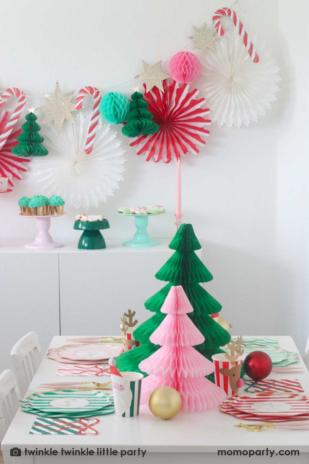A Christmas party table featuring Momo Party's festive striped party tableware in pink red and green with Christmas tree honeycombs as the centerpiece. In the back is a Christmas dessert table decorated with Momo Party's Christmas honeycomb decorations featuring candy canes, honeycomb Christmas trees and mint honeycomb balls. On the table there are multiple cake stands in red, pink, green and mint with Holiday treats like cookies, cupcakes and baked food, making this a perfect Holiday party inspo.