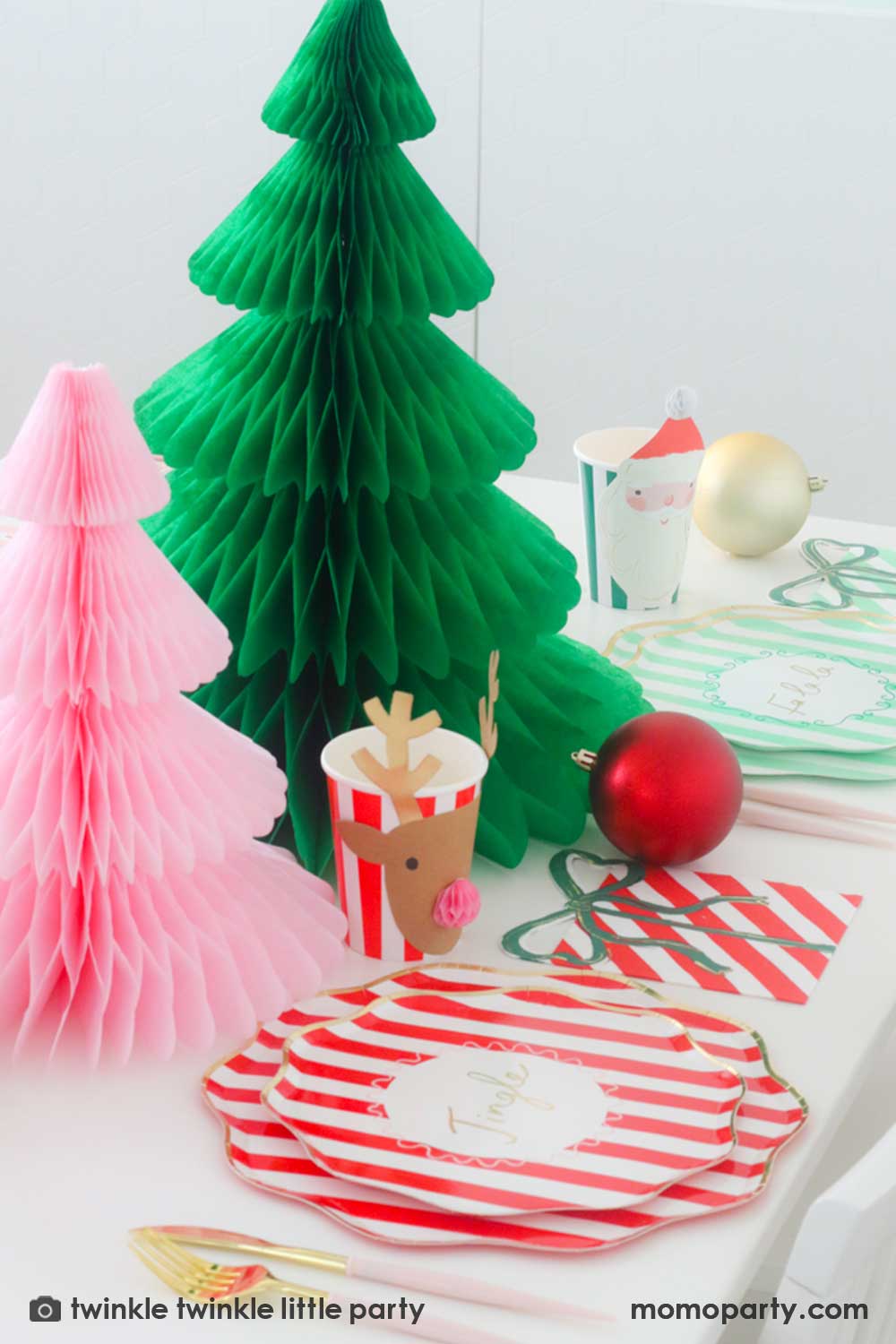 A festive Christmas party table featuring Momo Party's Festive striped dinner plates, side plates, present with a bow shaped napkins and Christmas honeycomb party cups in Santa design by Meri Meri. In the center of the table are a green and pink honeycomb Christmas tree decorations as the centerpiece. Along with some Christmas ornaments on the table, this makes a modern Holiday place setting inspo for a festive party this holiday season.