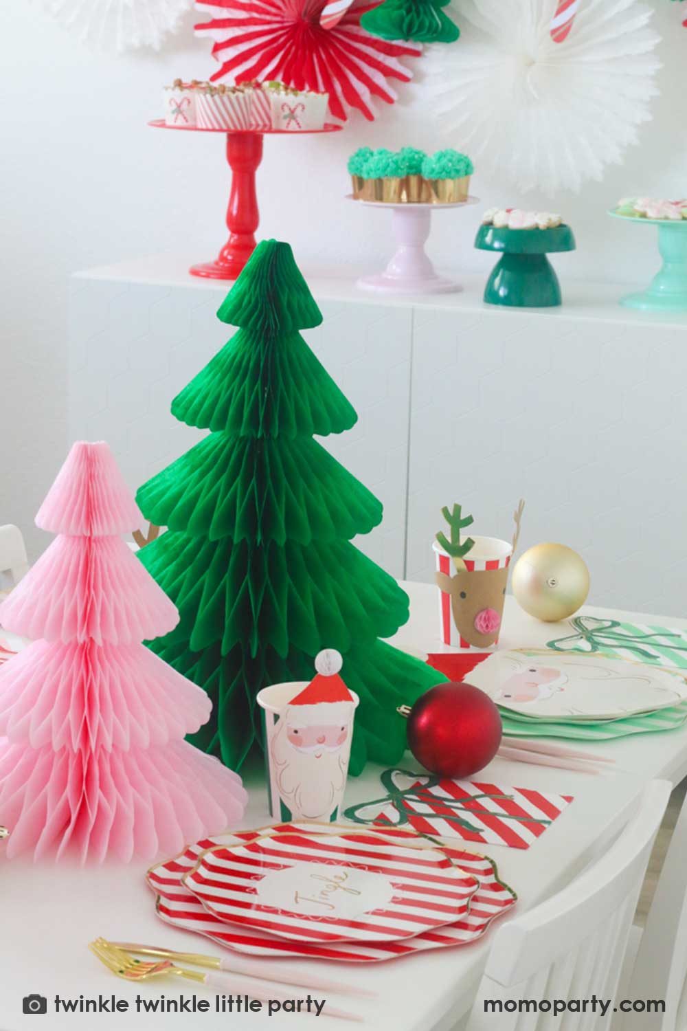 A festive Christmas party table featuring Momo Party's Festive striped dinner plates, side plates, present with a bow shaped napkins and Christmas honeycomb party cups in Santa design by Meri Meri. In the center of the table are a green and pink honeycomb Christmas tree decorations as the centerpiece. Along with some Christmas ornaments on the table, this makes a modern Holiday place setting inspo for a festive party this holiday season.