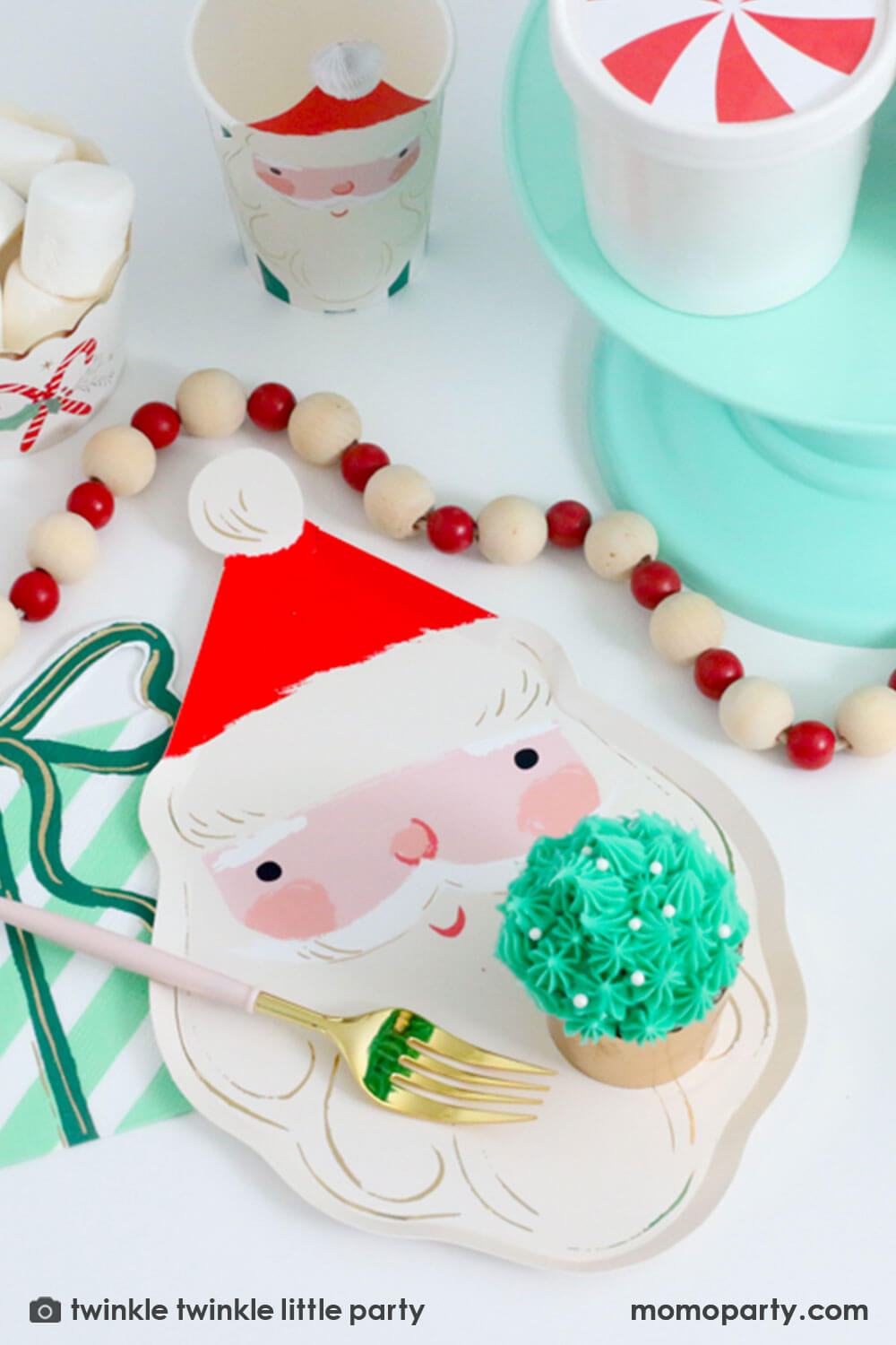 A festive Christmas tablescape featuring Momo Party's Jolly Santa Plate, present with a bow shaped napkin and Christmas honeycomb party cup in Santa design by Meri Meri. On the Jolly Santa plate there is a buttercream Christmas tree cupcake in festive gold foil baking cups and a festive red and white pom pom banner around the tablescape, making this a modern Holiday placeset inspo for a festive Holiday party.