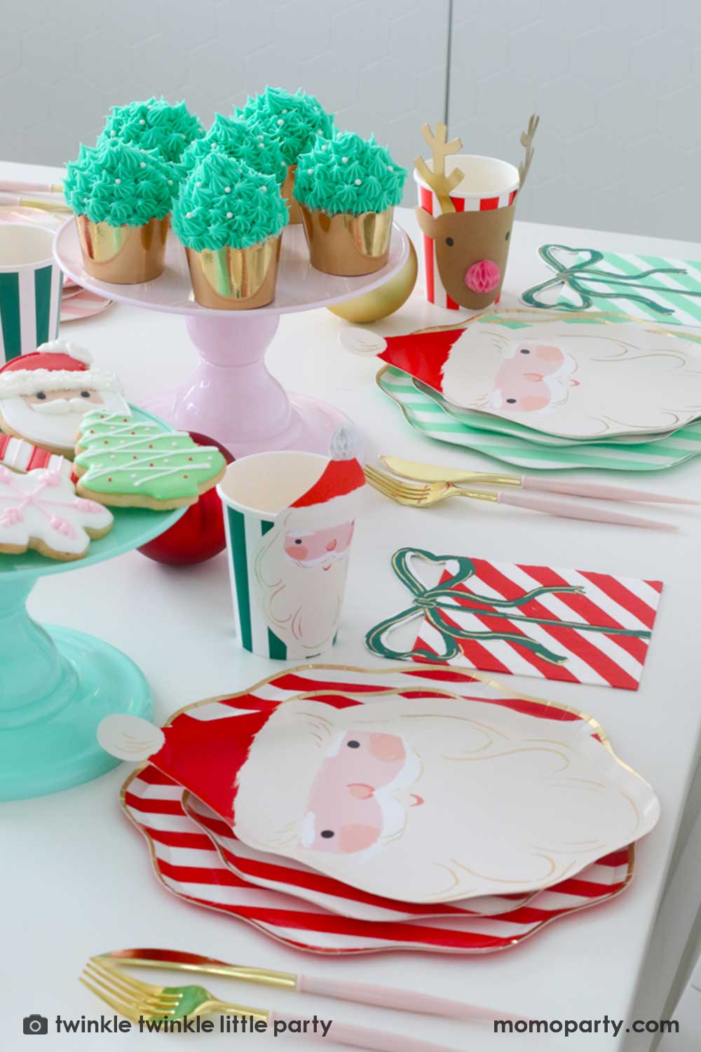 A festive Christmas party table featuring Momo Party's Festive striped dinner plates, side plates, Jolly Santa Plates, present with a bow napkins and Christmas honeycomb party cups by Meri Meri. On the center of the table are some buttercream Christmas tree cupcakes in festive gold foil baking cups and a mint cake topper with festive Holiday cookies in the design of a Santa, Christmas tree, candy cane and snowflake. Makes this a modern inspo for a festive Holiday table setting.