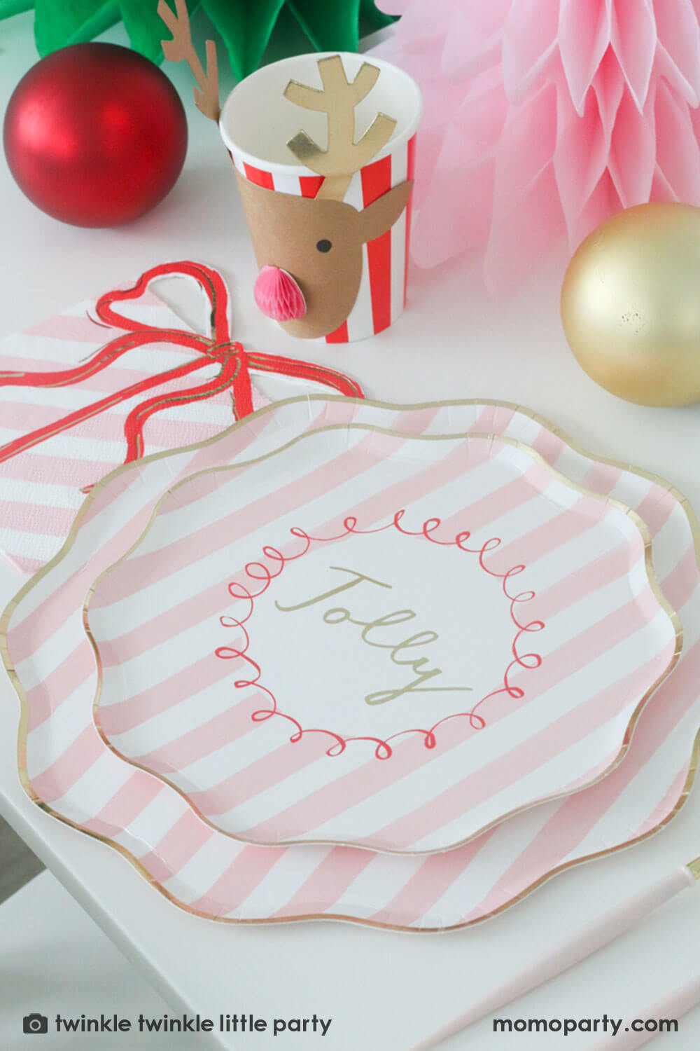 A festive Christmas party table featuring Momo Party's Festive pink striped dinner plate layered by a side plate and a pink present with a bow shaped napkin and a reindeer red honeycomb party cup by Meri Meri. In the center of the table are a green and pink honeycomb Christmas trees along with red and gold Christmas ornaments, making this a gorgeous Holiday tablescape for a darling Christmas party this holiday season.