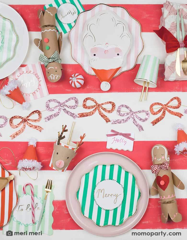 A festive Christmas party table featuring Momo Party's new Christmas party collection including festive striped plates, napkins and party cups in green, pink and red. On the red striped tablecover, there are also Meri Meri's Christmas honeycomb party cups in reindeer design, which paired with Santa shaped plates and glittering red and pink bows garland in the middle, makes it a festive and fun tablescape for kid's friendly Christmas party.