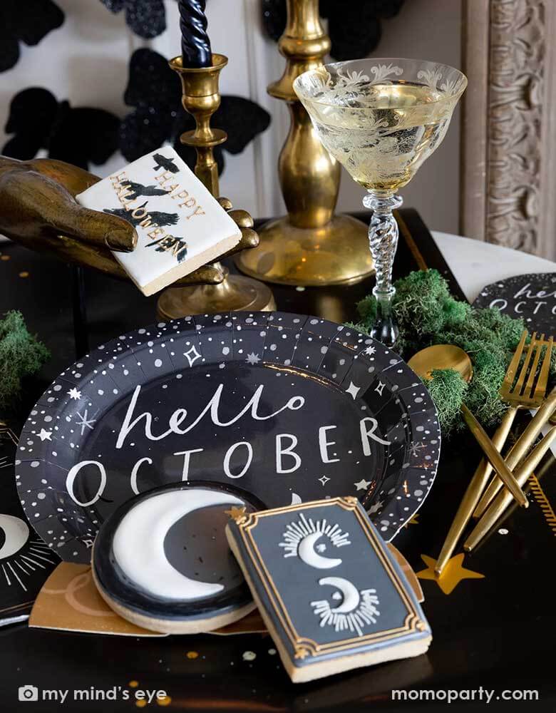 A mystical Halloween table decorated with Momo Party's moons and stars themed party supplies including a crystal ball shaped plate, moons and stars gold foil napkins, moons and stars table runner by My Mind's Eye. With gold metal candle stands holding black candles and vintage gold utensils with Halloween themed sugar cookies and black glittered butterfly decorations on the wall, all makes a perfect inspo for an elegant and chic wizard and witch themed Halloween party decorating idea.