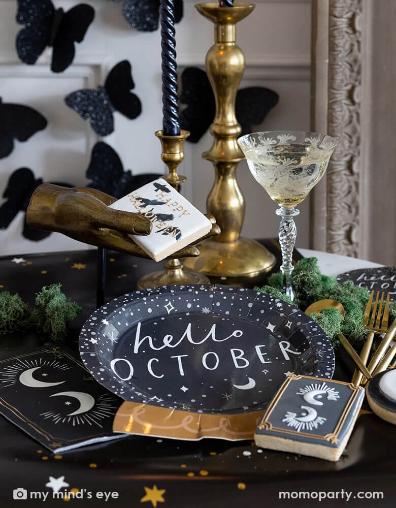 A mystical Halloween table decorated with Momo Party's moons and stars themed party supplies including a crystal ball shaped plate, moons and stars gold foil napkins, moons and stars table runner by My Mind's Eye. With gold metal candle stands holding black candles and vintage gold utensils with Halloween themed sugar cookies and black glittered butterfly decorations on the wall, all makes a perfect inspo for an elegant and chic wizard and witch themed Halloween party decorating idea.